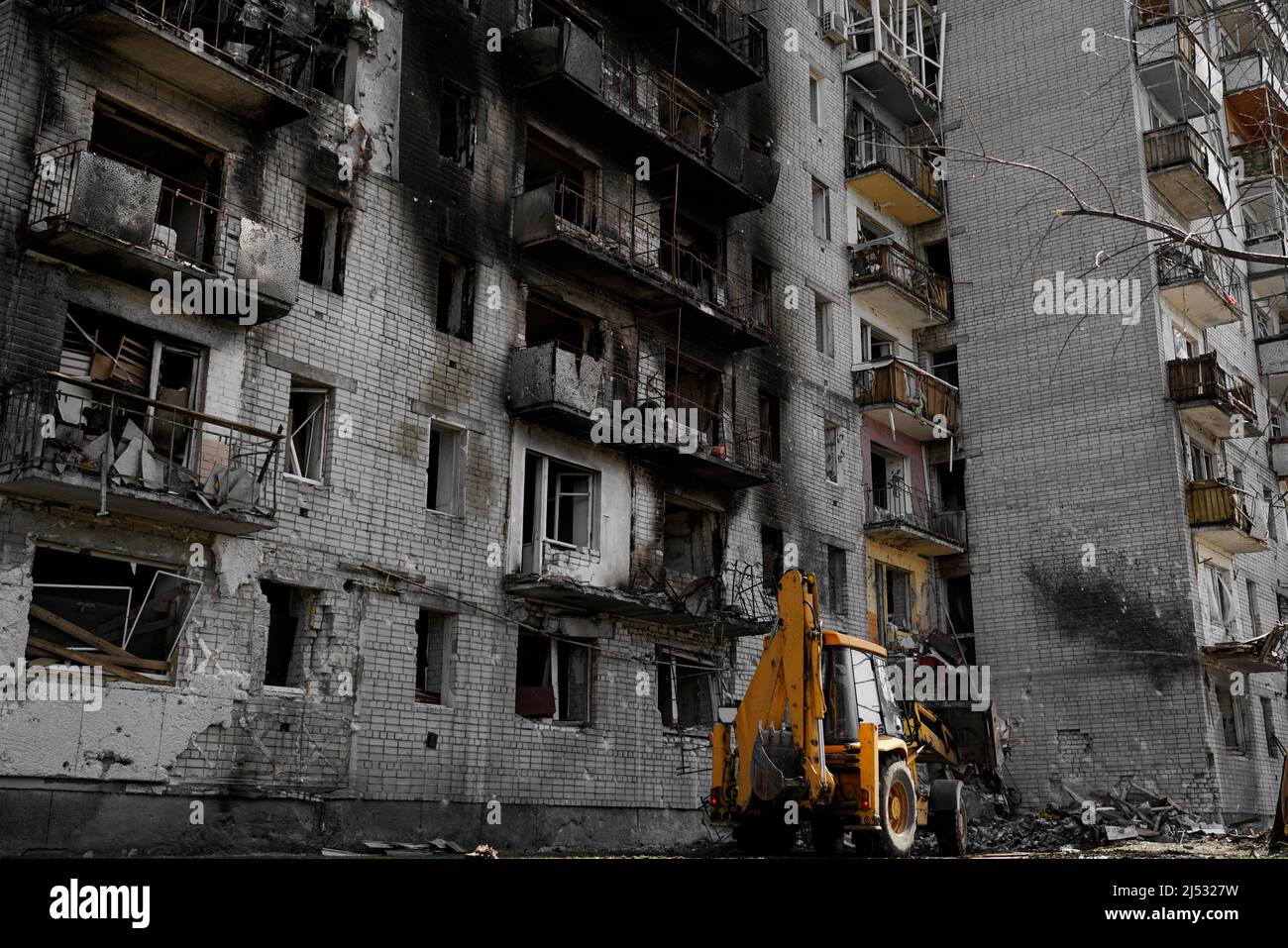 Ein verbranntes, zerstörtes Wohnhaus ohne Fenster, das während des Beschuss und der Militäroperationen in der Ukraine beschädigt wurde. Unter dem Haus räumt der Traktor Stockfoto