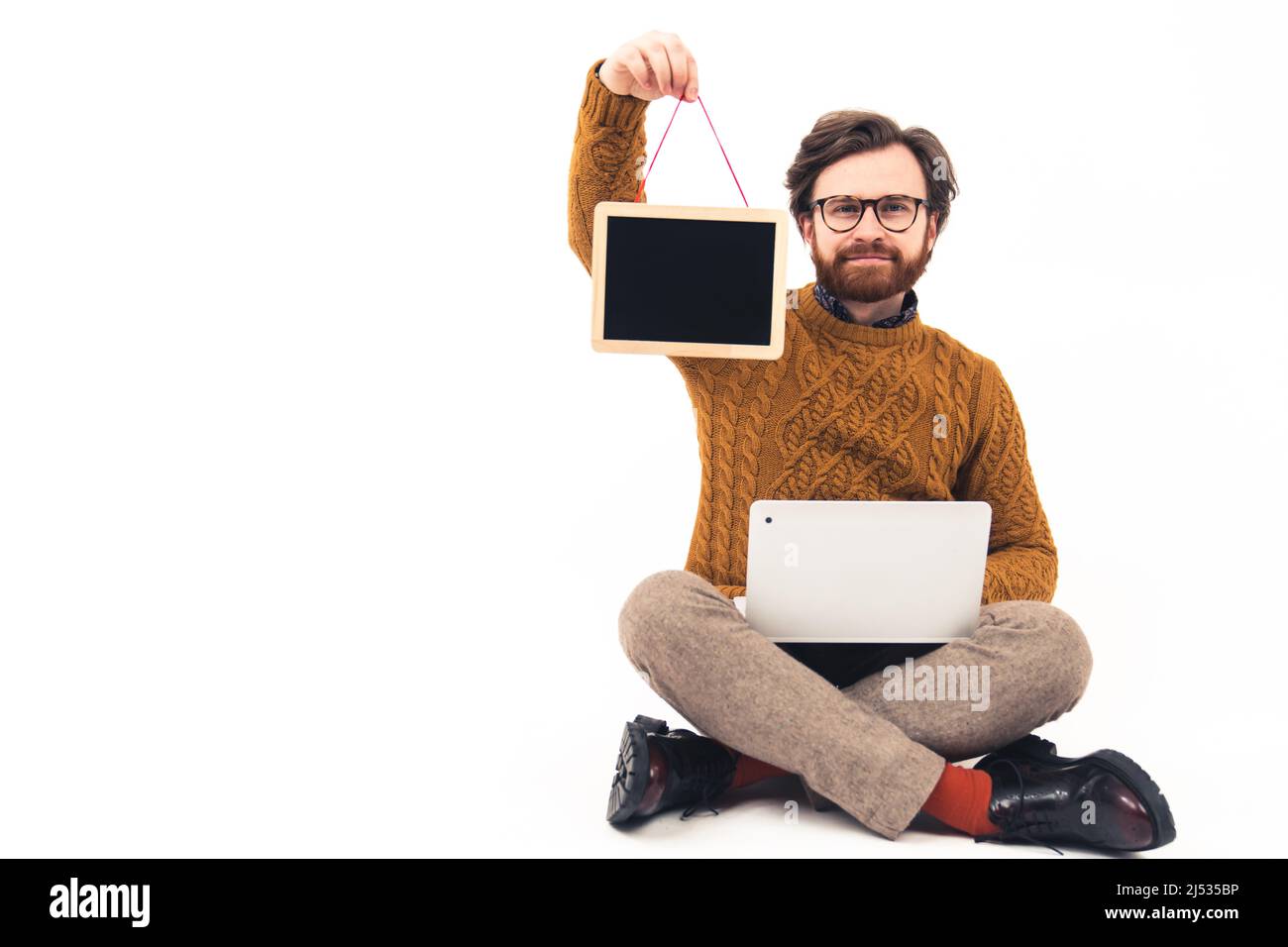 Bärtiger Mann, der mit gekreuzten Beinen sitzt und einen Laptop auf den Knien hält und einen kleinen schwarzen Schreibtisch an einer Schnur zeigt. Hochwertige Fotos Stockfoto