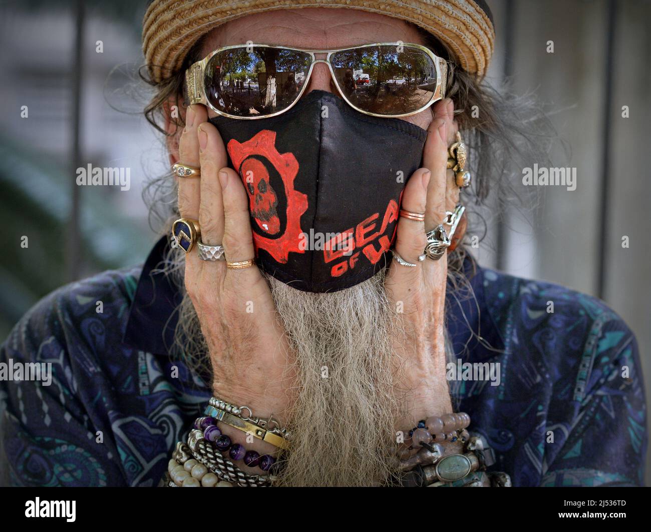Älterer kaukasischer Mann mit verspiegelter dunkler Sonnenbrille und handbemalter schwarzer Stoffmaske (Gear of war) legt seine beringten Hände auf seine Wangen. Stockfoto