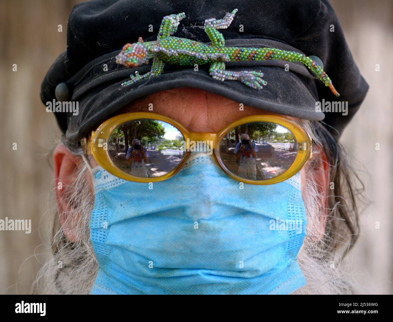 Das Porträt eines älteren kaukasischen Mannes mit verspiegelter Sonnenbrille und hellblauer chirurgischer Gesichtsmaske trägt eine Spitzenkappe mit einem Reptilienabzeichen (Leguan). Stockfoto