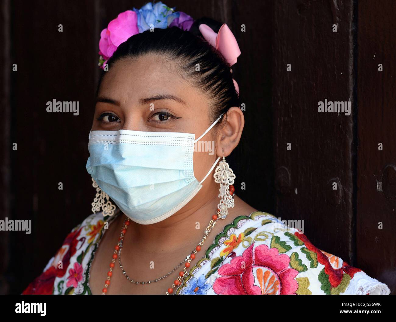 Die körperpositive junge mexikanische Frau mit Blumen in gesenktem Haar trägt ein farbenfrohes, traditionelles Yucatan Maya Huipil-Kleid und eine chirurgische Gesichtsmaske. Stockfoto