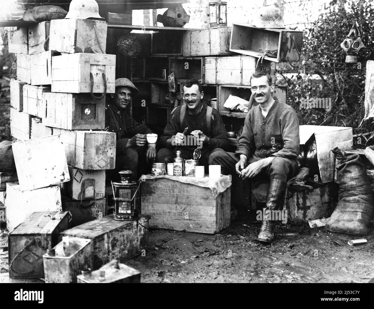 Drei Männer der Militärpolizei halten für etwas zu essen und zu trinken. Sie sitzen am Eingang eines aus Holzkisten gebauten Schutzheims. Die Kisten dienen als Regale, und einer wird als Tisch verwendet. Stockfoto
