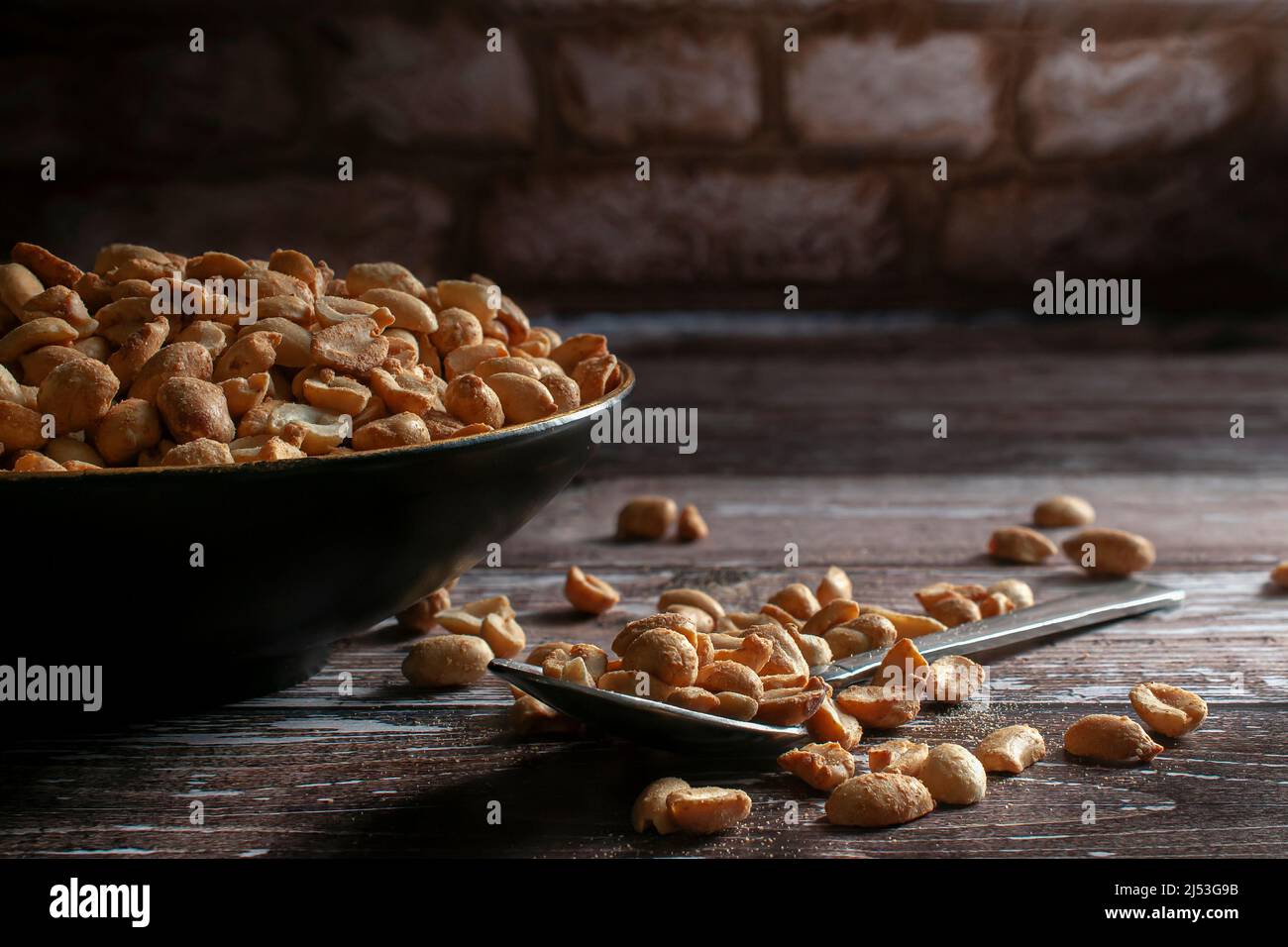 Geröstete Erdnüsse auf einer schwarzen Schüssel und auf einem Löffel über einem rustikalen Holztisch mit einer Ziegelwand im Hintergrund Stockfoto