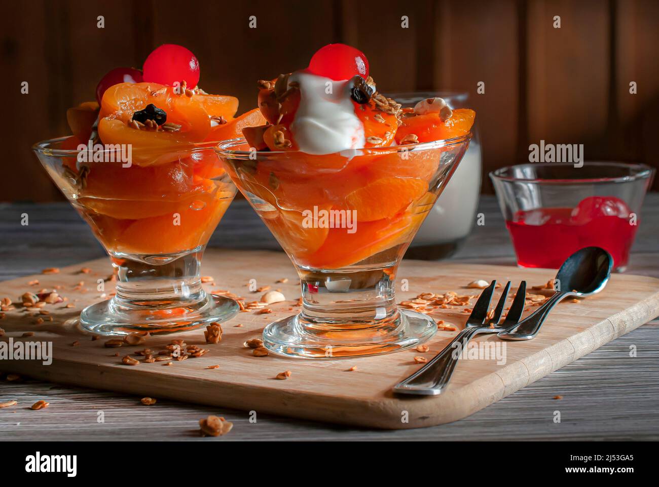 Zwei Glasbecher mit Aprikosenscheiben, Müsli-Joghurt und Kirschen auf der Oberseite, mit Besteck an der Seite und auf einem Schneidebrett, über einem Holztisch. Stockfoto