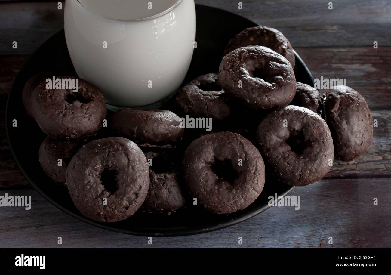 Ein schwarzer Teller mit kleinen Baby-Schokoladendonuts, die vom Morgenlicht beleuchtet werden, mit einer Eismilch an der Seite und auf einem Holztisch. Stockfoto