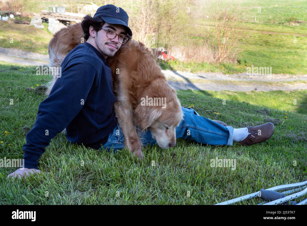 Ein hübscher Kaukasusmann sitzt allein auf einem grasbewachsenen Hügel mit Naturhintergrund und ein liebevoller goldener Retriever-Hund auf seinem Schoß mit einem Paar Krücken auf dem Schoß Stockfoto