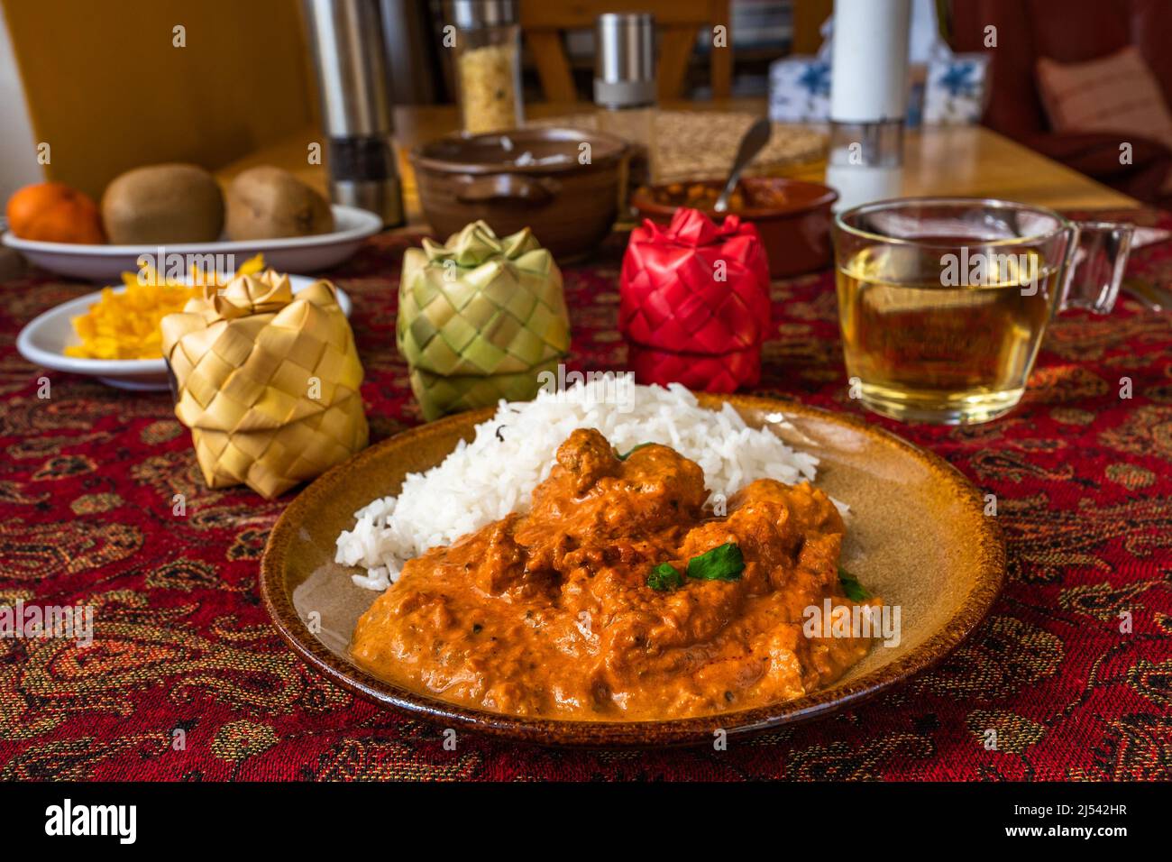 Würziges indisches Hühnchen Tikka Masala mit Reis auf Teller, 3 Bambuswürzkiste, Tasse Tee, Obst auf rotem Tischtuch auf Tisch. Stockfoto