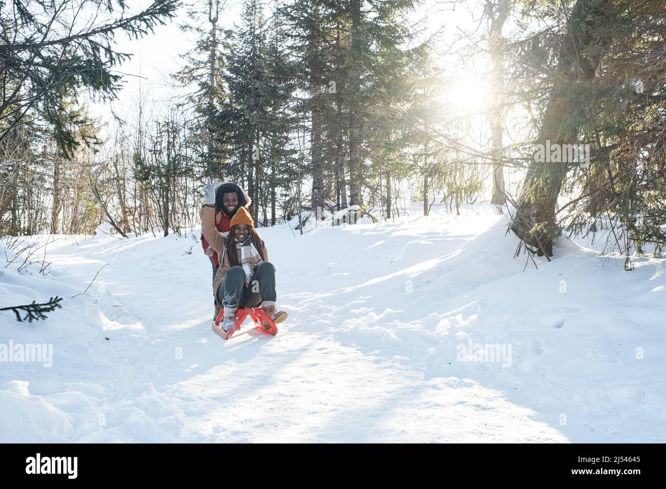 Lange Aufnahme des verspielten jungen schwarzen Mannes, der seine fröhliche Freundin am sonnigen Wintertag auf dem Schlitten im Wald schiebt Stockfoto
