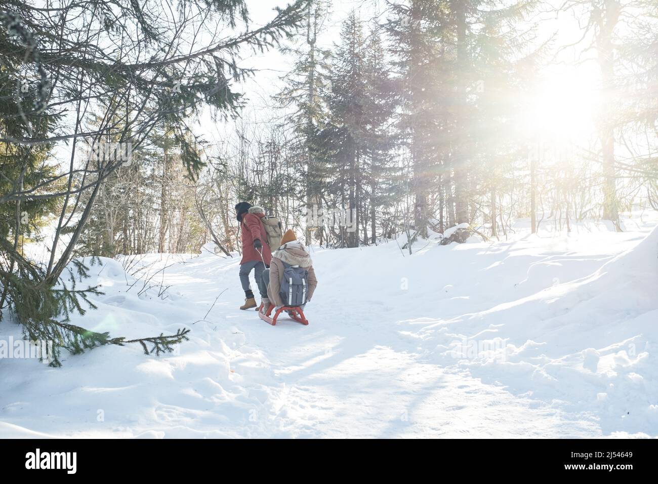 Lange Aufnahme eines jungen Mannes, der seine Freundin am sonnigen Wintertag im Wald in den Bergen auf dem Schlitten zieht Stockfoto