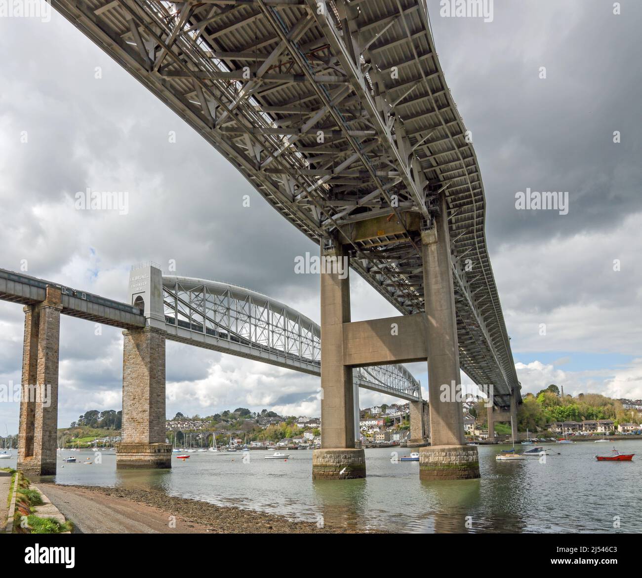 Ein Zug, der die Royal Albert Bridge vom Flussufer aus unter der Tamar Road Bridge überquert. Die Eisenbahnbrücke ist eine eingleisige historische Eisenbahn b Stockfoto