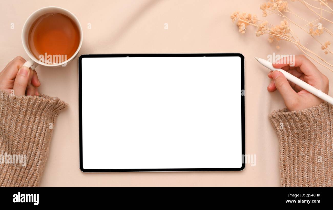 Aufnahme über den Kopf, Bloggerin oder Freiberuflerin mit einer Tasse Tee, während sie in ihrem minimalistischen Arbeitsbereich ein digitales Tablet verwendet. Modell mit weißem Bildschirm des Tablets. Stockfoto