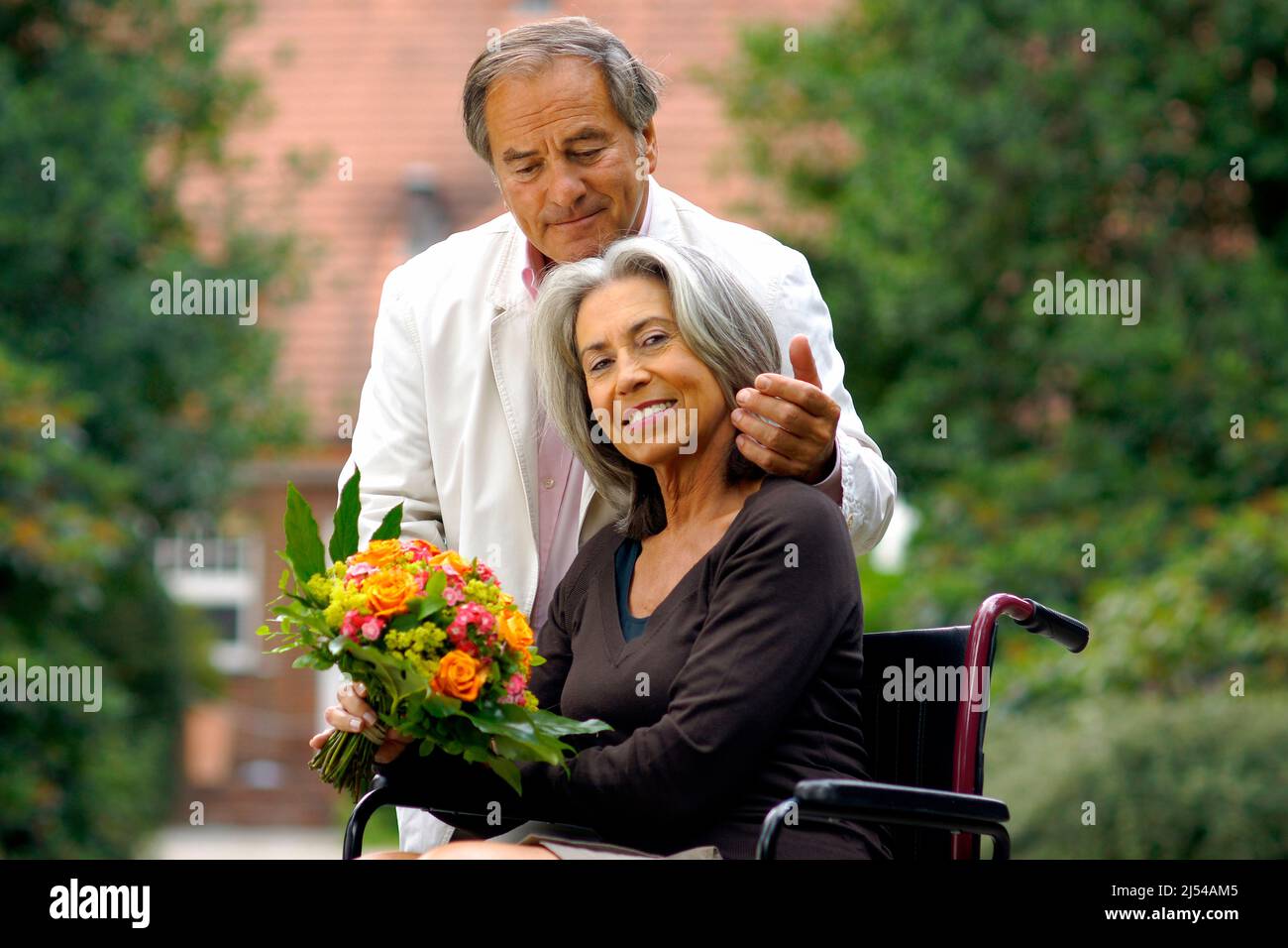 Ältere Paare in der Natur, die Frau sitzt in einem Rollstuhl mit einem Blumenstrauß und der Mann umarmt sie liebevoll Stockfoto