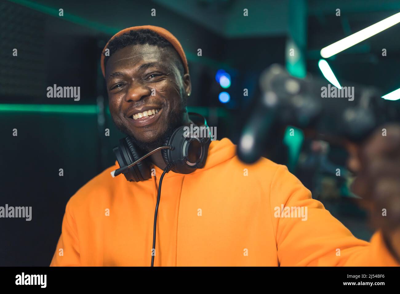 Porträt eines jungen lächelnden Afro-amerikanischen männlichen Spielers in orange Hoodie trägt Kopfhörer und halten Joystick dunklen Hintergrund copy Space . Hochwertige Fotos Stockfoto
