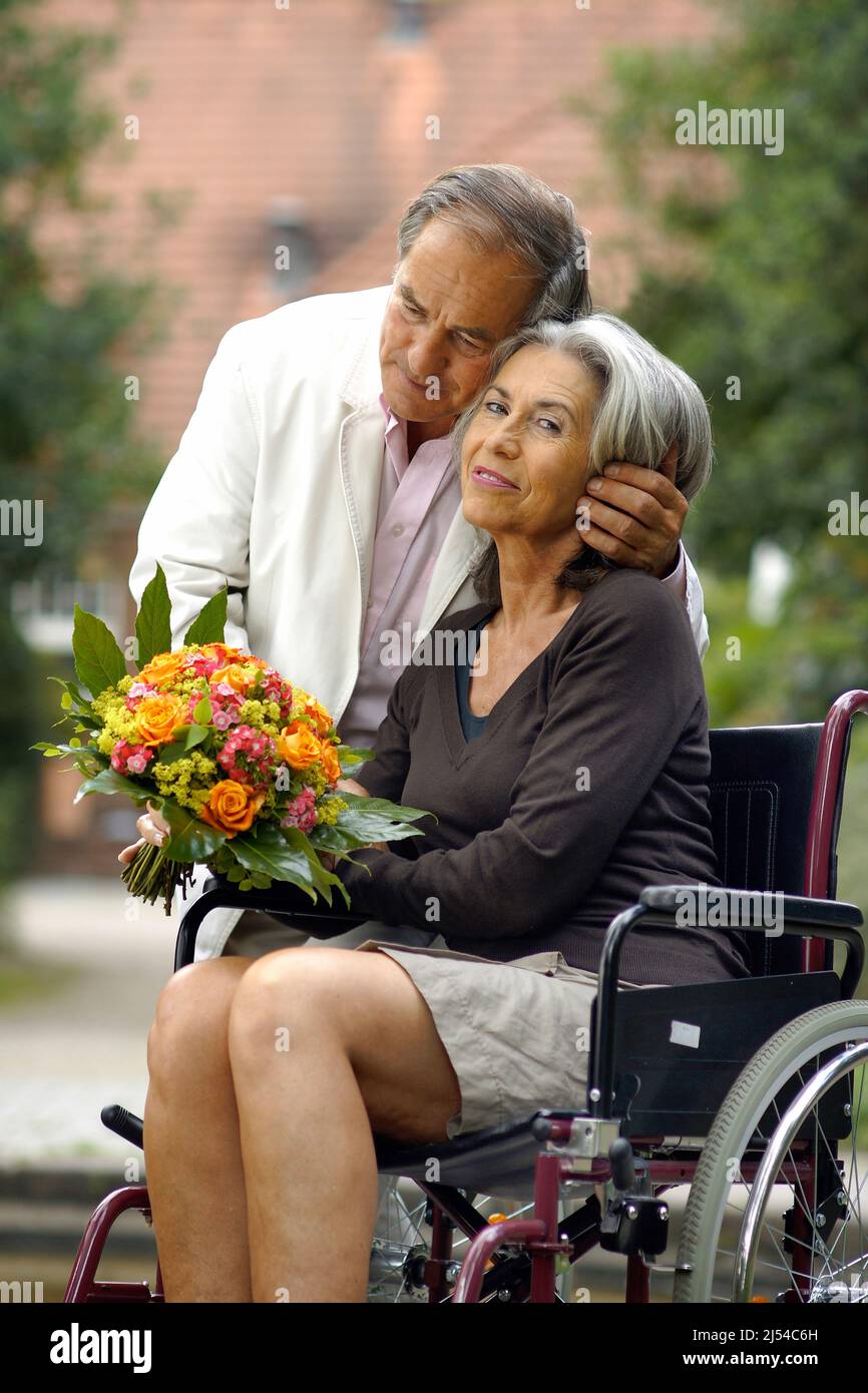 Ältere Paare in der Natur, die Frau sitzt in einem Rollstuhl mit einem Blumenstrauß und der Mann umarmt sie liebevoll Stockfoto