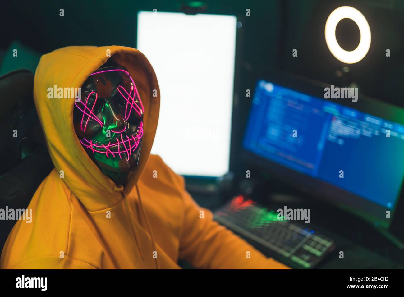Anonymes Internet-Identitätskonzept. Person in einem gelben Hoodie vor einem Programmiercomputer mit gruselig beleuchteter Neonmaske und Blick auf die Kamera. Hochwertige Fotos Stockfoto