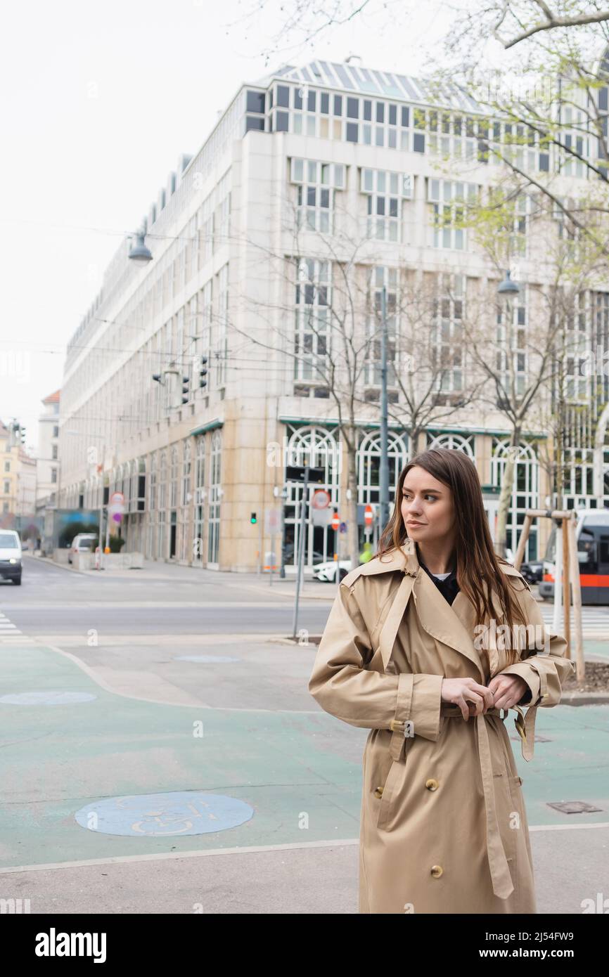 Junge Frau, die den Gürtel auf einem stylischen Trenchcoat auf der Straße der europäischen Stadt anpasst Stockfoto