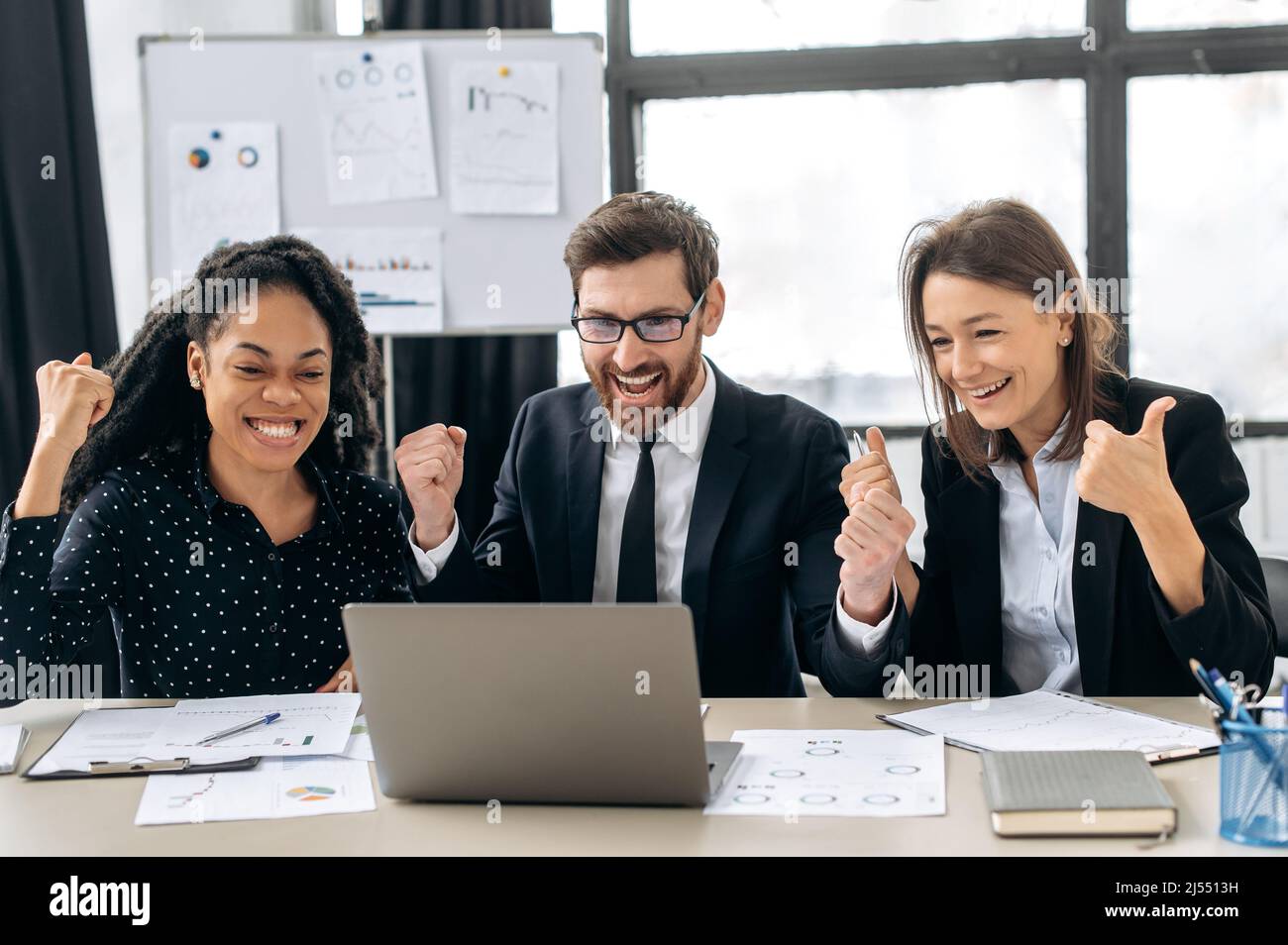 Sieg, Sieg. Fröhliche kreative Geschäftspartner, die zusammenarbeiten. Multirassische Kollegen denken gemeinsam an Strategien, sitzen im modernen Büro, nutzen Laptops, bekommen gute Angebote, profitieren, lächeln Stockfoto