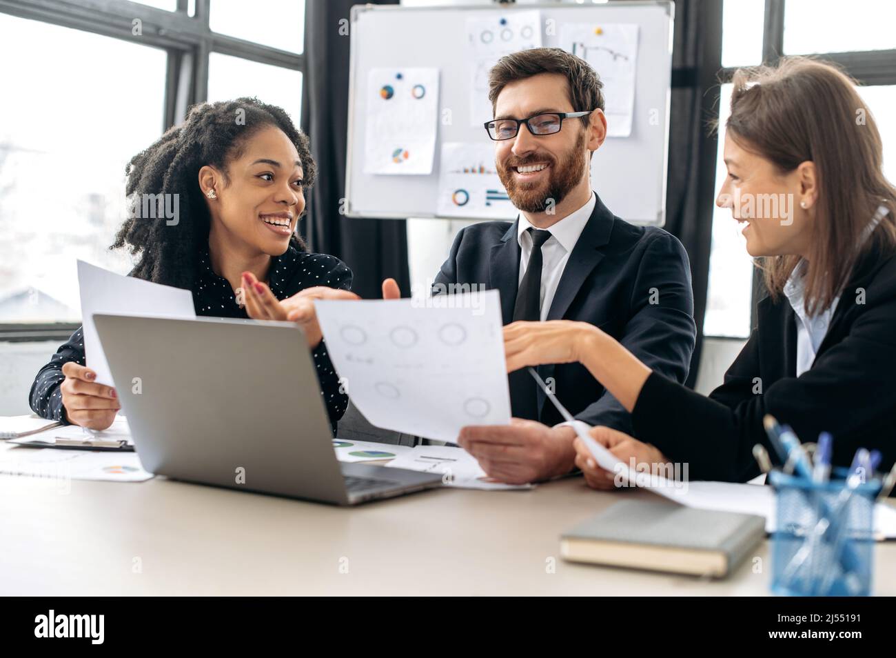 Erfolgreiche kreative Geschäftspartner, die an einem Projekt arbeiten. Multirassische Kollegen denken gemeinsam an Strategien, sitzen im modernen Büro, studieren Dokumente, benutzen Laptops, diskutieren, lächeln Stockfoto