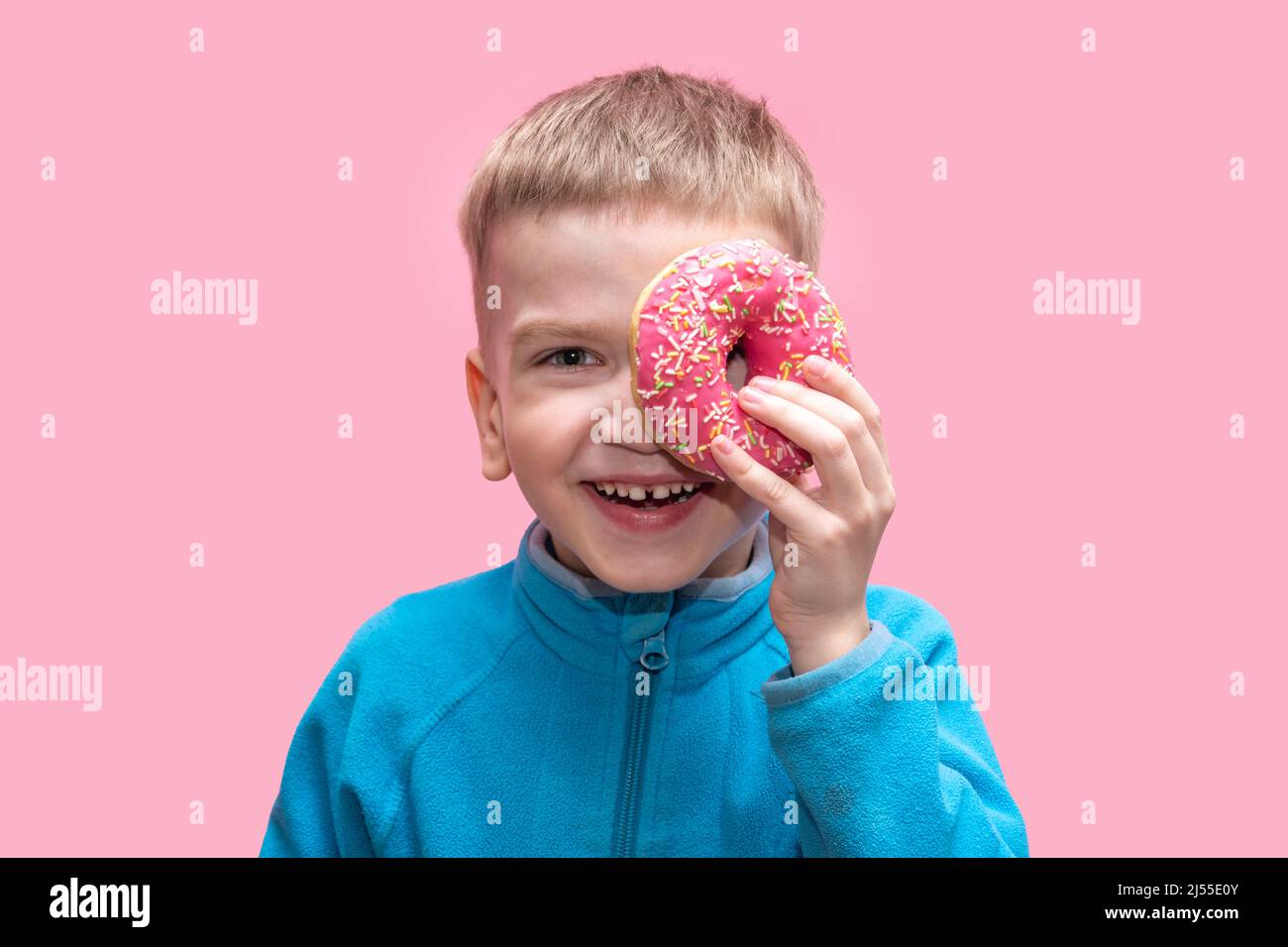 Ein süßer, lustiger Junge in einem blauen Pullover hält einen leuchtend rosa Donut in der Nähe seines Auges und lacht auf einem rosa Hintergrund. Liebenswert fröhlich Kind spielt mit einem don Stockfoto