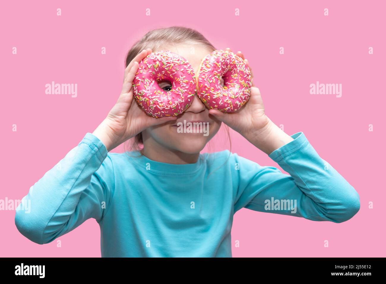 Ein niedliches lustiges Mädchen in einem blauen T-Shirt hält leuchtend rosa Donuts wie eine Brille in der Nähe ihrer Augen und sieht dich auf einem rosa Hintergrund an. Fröhliche Kindheit Konz Stockfoto