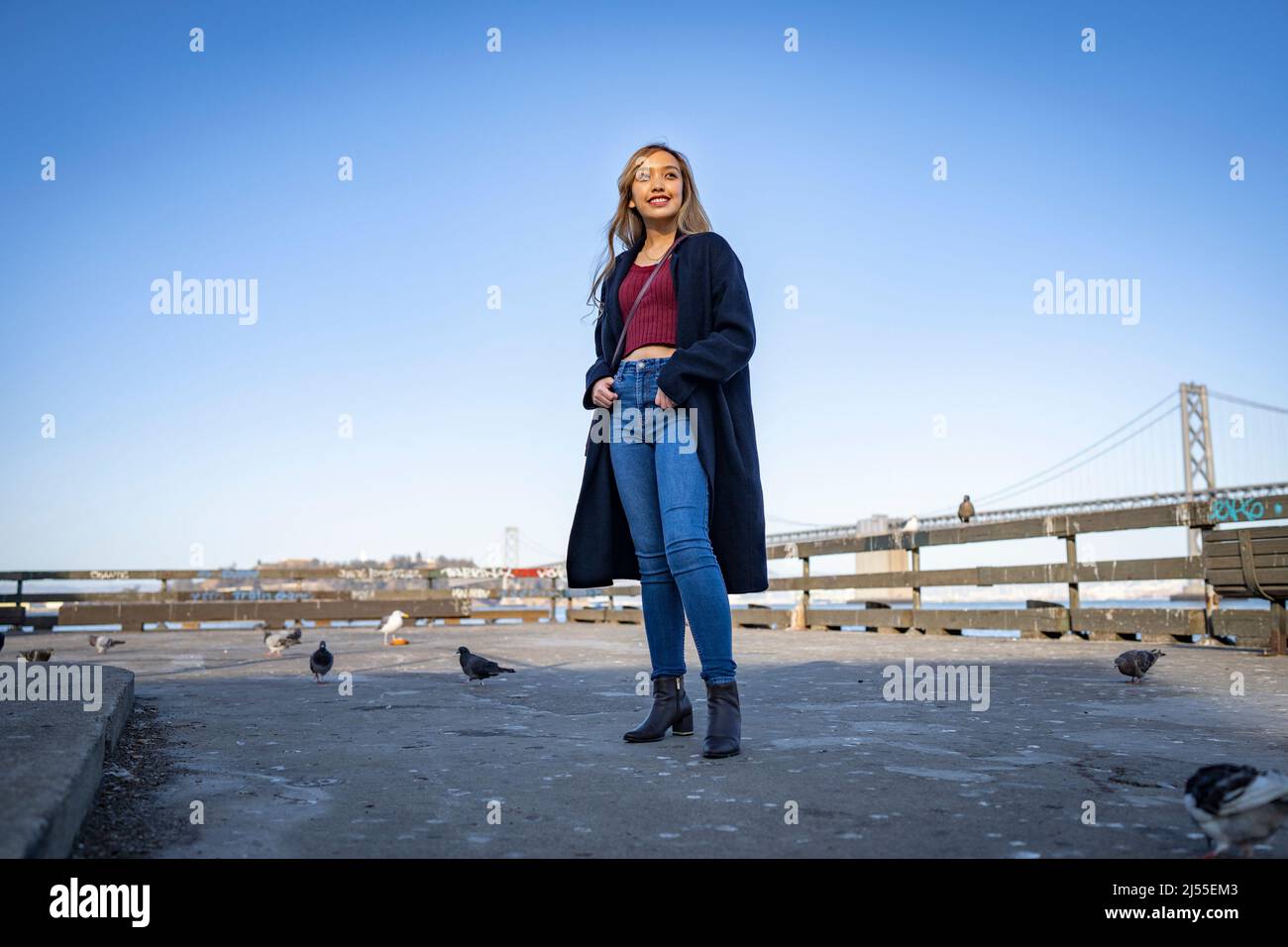 Lächelnde/glückliche junge Frau am Pier mit der Oakland Bay Bridge im Hintergrund | Lifestyle Local Tourism Stockfoto