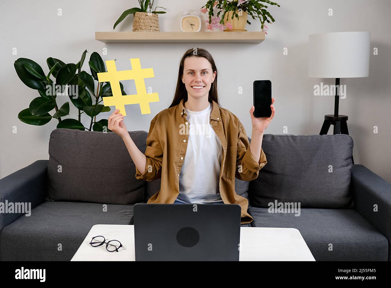 Lächelnd attraktive junge Bloggerin, die zu Hause auf einem bequemen Sofa sitzt und ein großes gelbes Hashtag-Symbol und ein Smartphone zeigt, glücklich in die Kamera schaut, Stockfoto