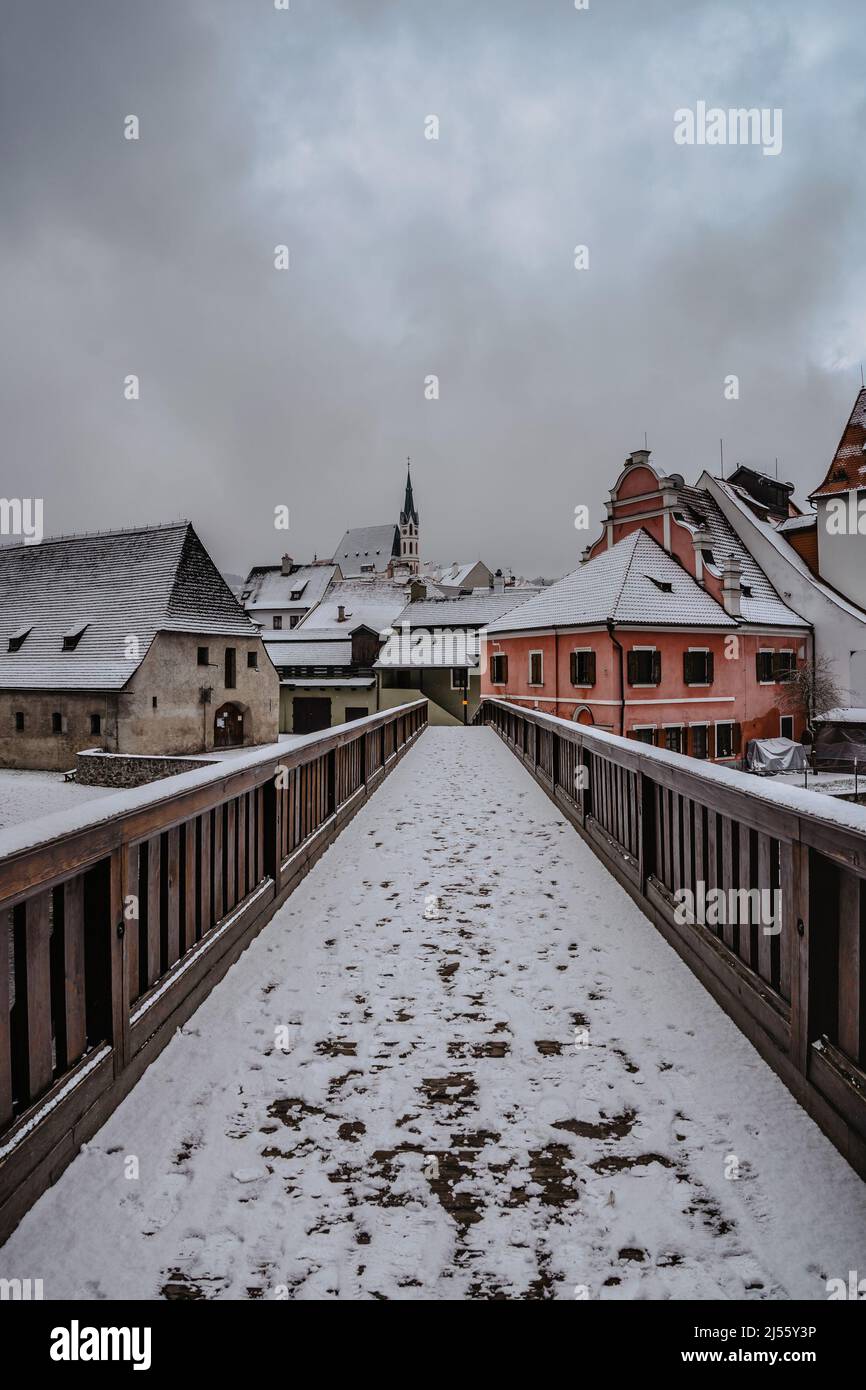 Schneebedeckter Holzweg über den Fluss Moldau, Gebäude und Kirche im Hintergrund, Cesky Krumlov, Tschechische republik. Winterlicher Tag, leere Straße ohne Touris Stockfoto