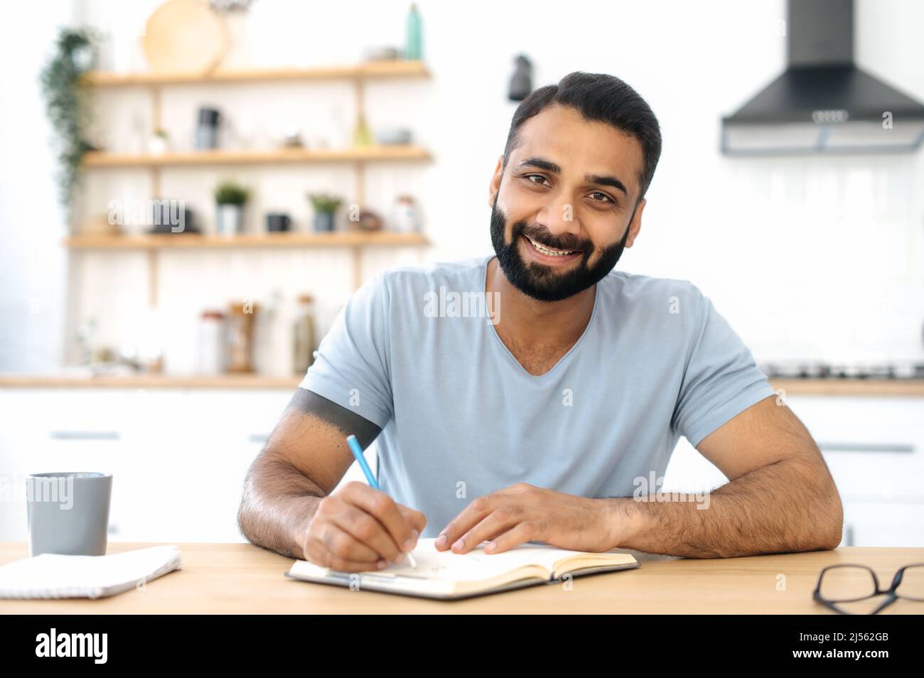 Ein intelligenter, positiver Inder, der zu Hause an einem Tisch in der Küche sitzt, einem Vortrag zuhört, per Videokonferenz ein Webinar abhält und sich Notizen in einem Notizbuch macht. Fernunterricht, Weiterbildung Stockfoto