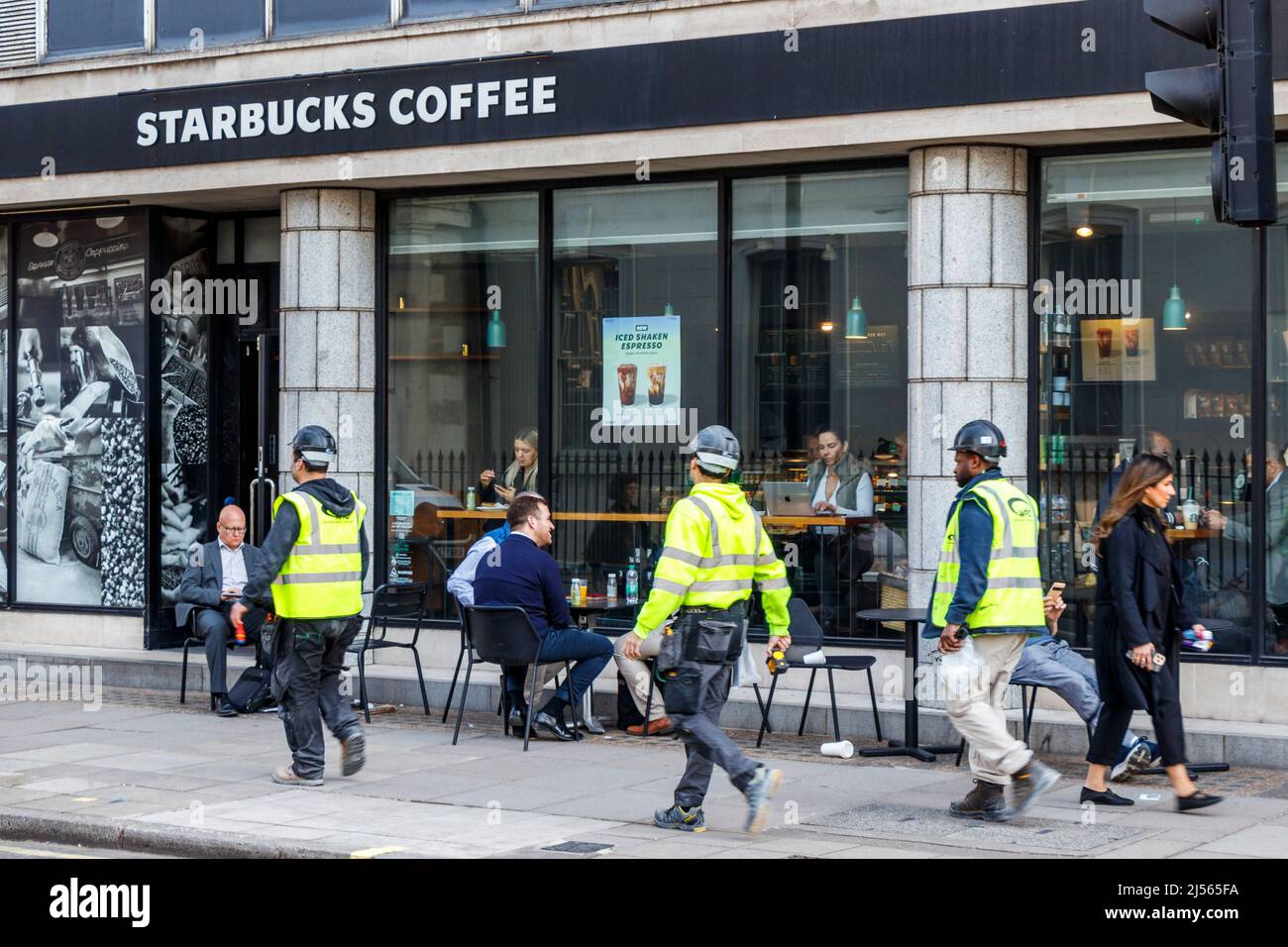 Ein Trio von Arbeitern in Jacken mit hohen Mänteln geht an Gästen vorbei, die vor einer Filiale von Starbucks in der Margaret Street, London, sitzen, während das Wetter in Großbritannien wärmer wird Stockfoto