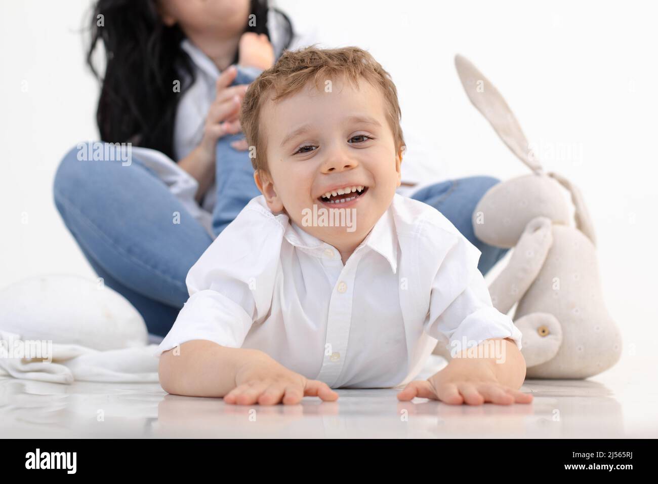 Foto eines kleinen, hellhaarigen Jungen, der auf dem Boden liegt, in der Nähe eines weichen Spielzeughasen. Blick auf die Kamera, spielt mit der Mutter zusammen Stockfoto