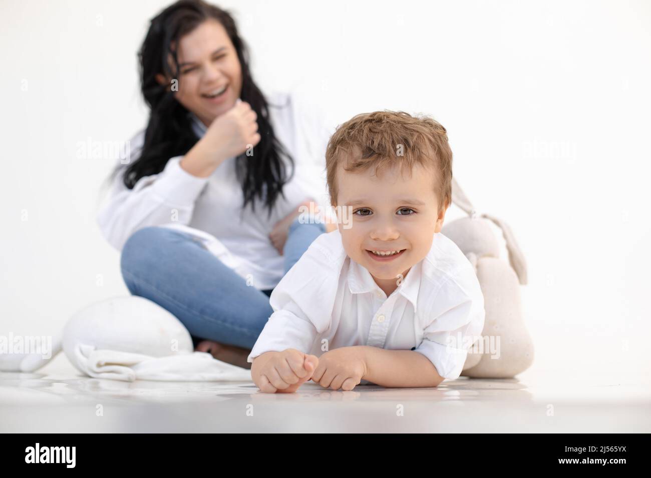 Foto eines kleinen, hellhaarigen Jungen, der auf dem Boden liegt, in der Nähe eines weichen Spielzeughasen. Kamera betrachten, spielen und verspotten, Freizeit Stockfoto