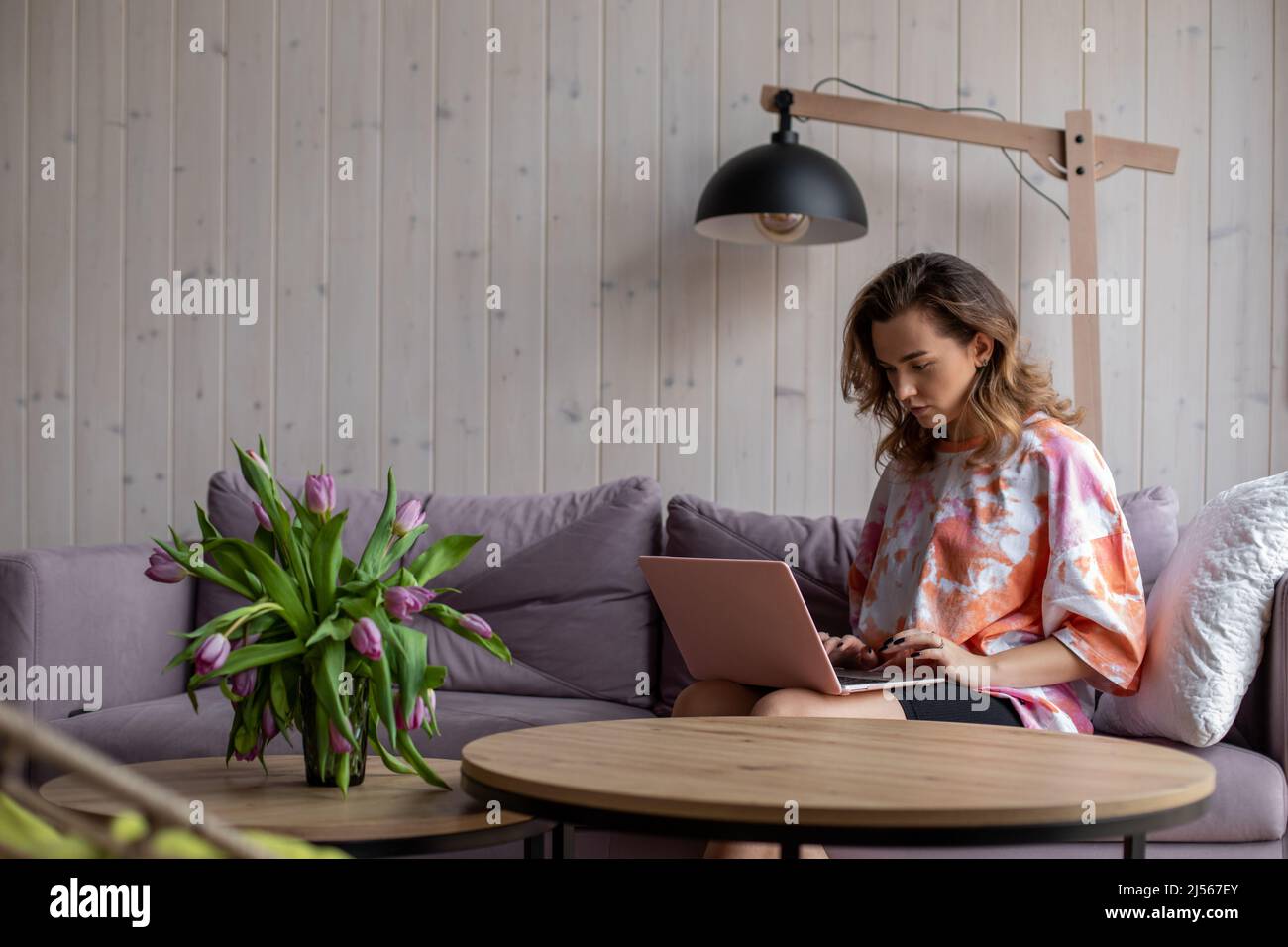 Porträt einer Frau, die auf der Tastatur des Laptops tippt und auf einem weichen Sofa in legerer Kleidung neben dem Tisch sitzt, mit einer Vase mit ausbleichenden Tulpen. Wohnzimmer in Stockfoto