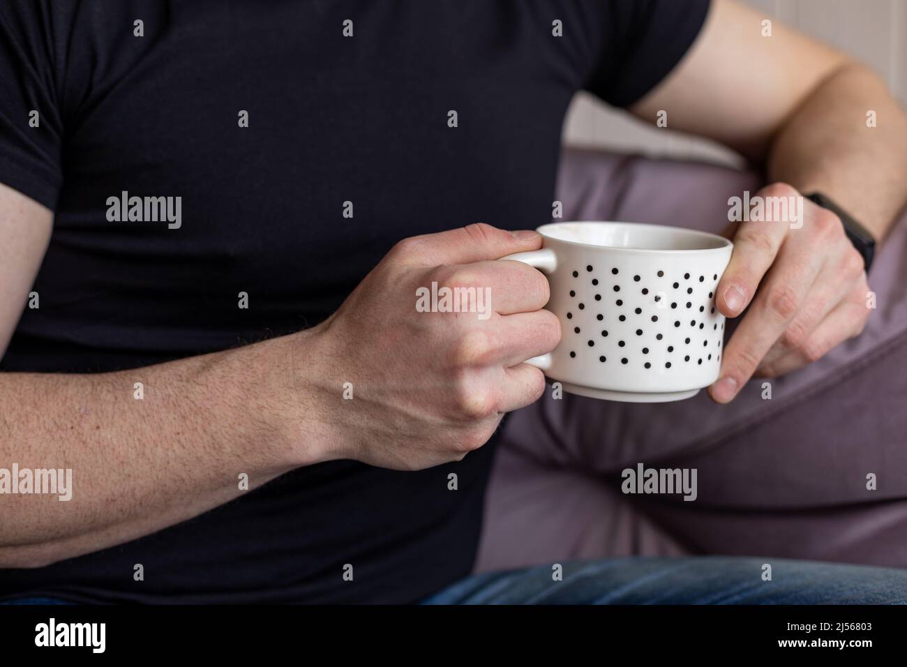 Porträt eines Mannes, der eine weiße Tasse Kaffee in der Hand hält und auf einem weichen, violetten Sofa in legerer Kleidung in Nahaufnahme sitzt, verschwommener Hintergrund. Wohnzimmer in elegant Stockfoto