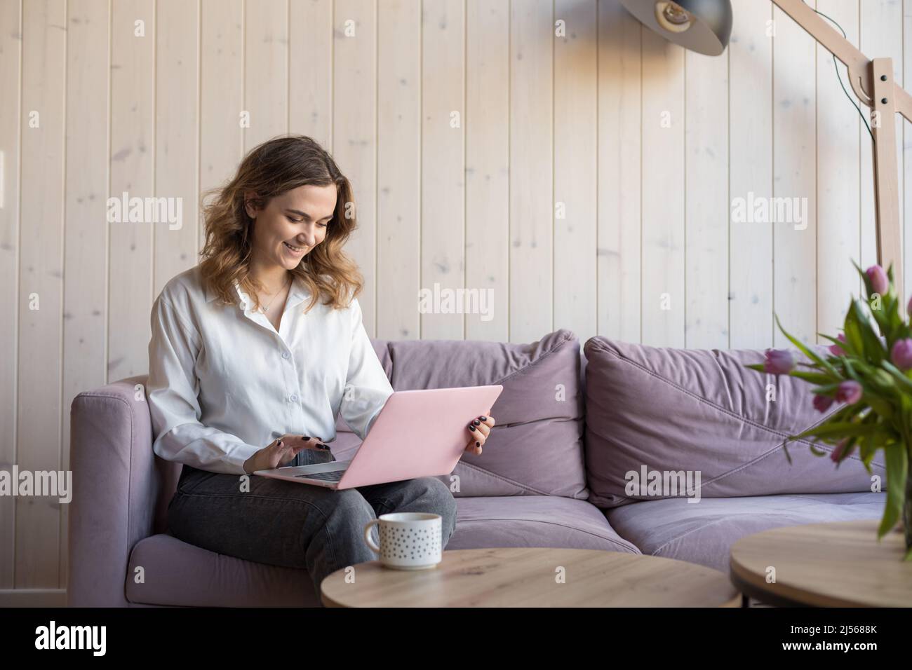 Porträt einer lächelnden Frau, die auf einem rosa Laptop auf einem weichen violetten Sofa in legerer Kleidung neben dem Tisch mit einer Tasse Kaffee sitzt. Wohnzimmer in elegant Stockfoto