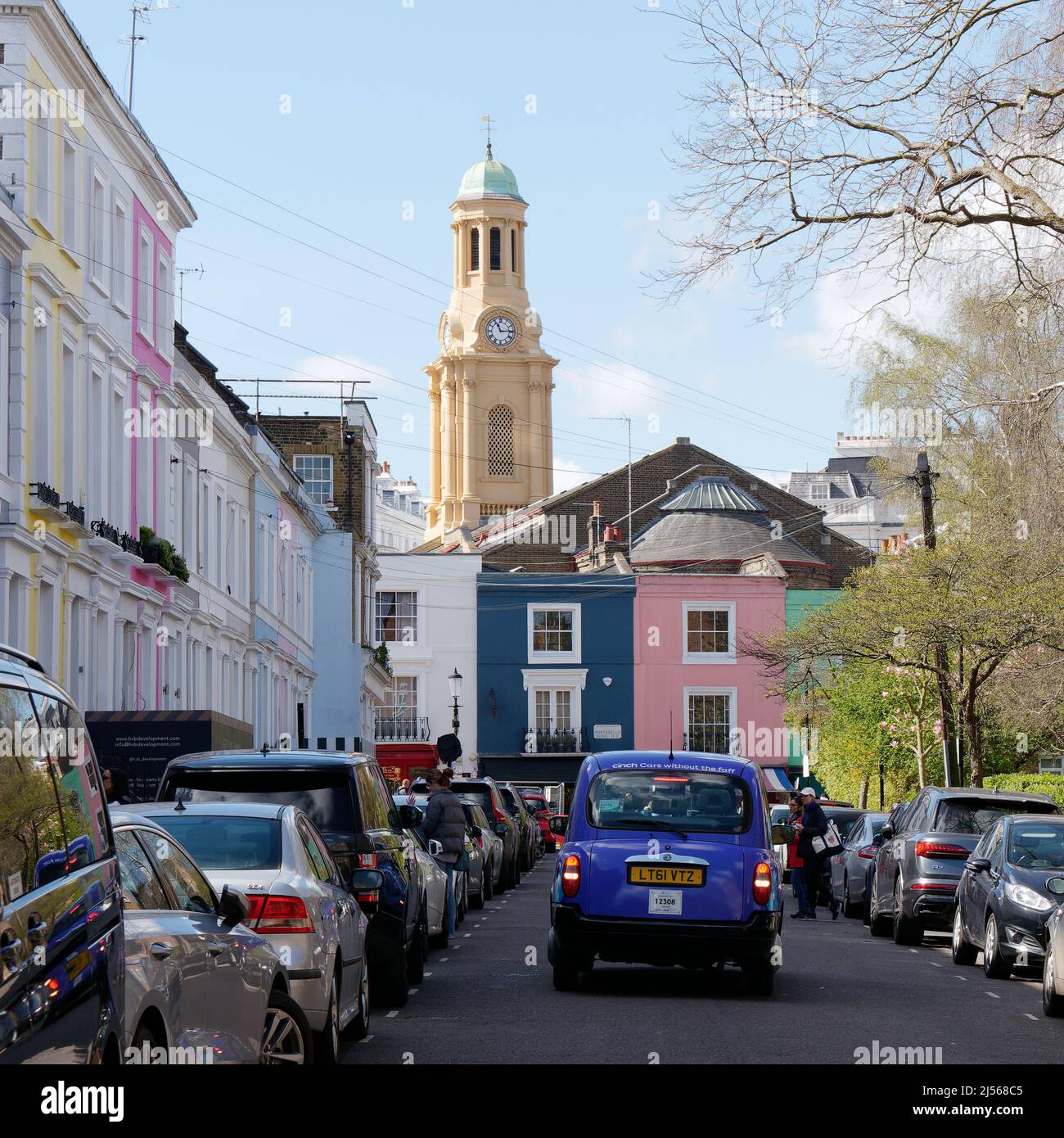 London, Greater London, England, April 09 2022: Farbenfrohe Anwesen in Notting Hill und ein lila Taxi mit dem Glockenturm der St. Peters Kirche dahinter Stockfoto