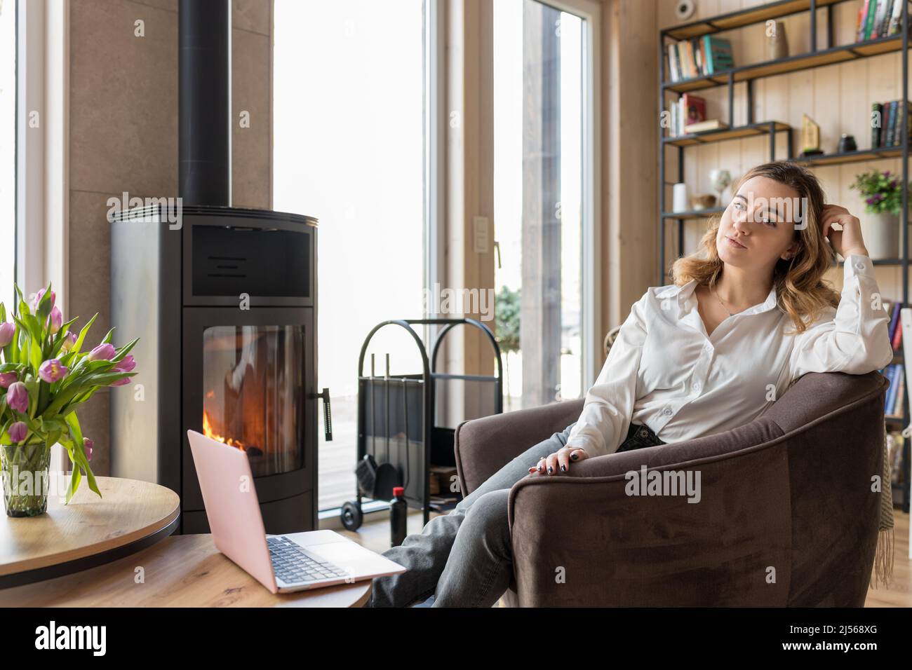 Porträt einer glücklichen Frau mit Laptop, die in legerer Kleidung neben Fenster und Kamin auf einem weichen Sessel sitzt. Wohnzimmer in elegantem Design mit Ästhetik Stockfoto