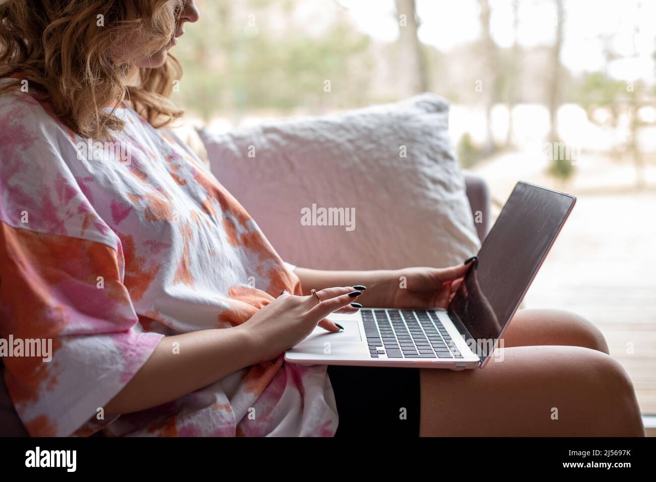 Nahaufnahme einer jungen Frau, die auf einem modernen Laptop tippt und arbeitet und auf einem weichen Sofa in legerer Kleidung in der Nähe eines großen Panoramafensters sitzt. Wohnzimmer Stockfoto