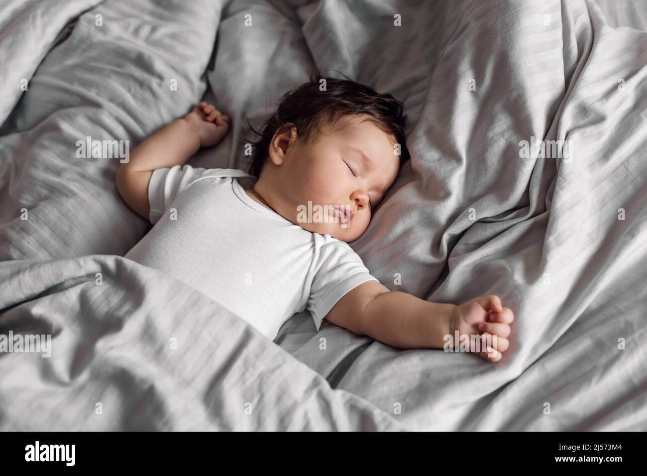 Porträt eines Babys in Schlafkleidung, das zu Hause unter der Decke auf dem Bett liegt. Schlafende Hände auseinander Kleinkind im Schlafzimmer, siehe süßen Traum. Grauer Hintergrund, frei Stockfoto