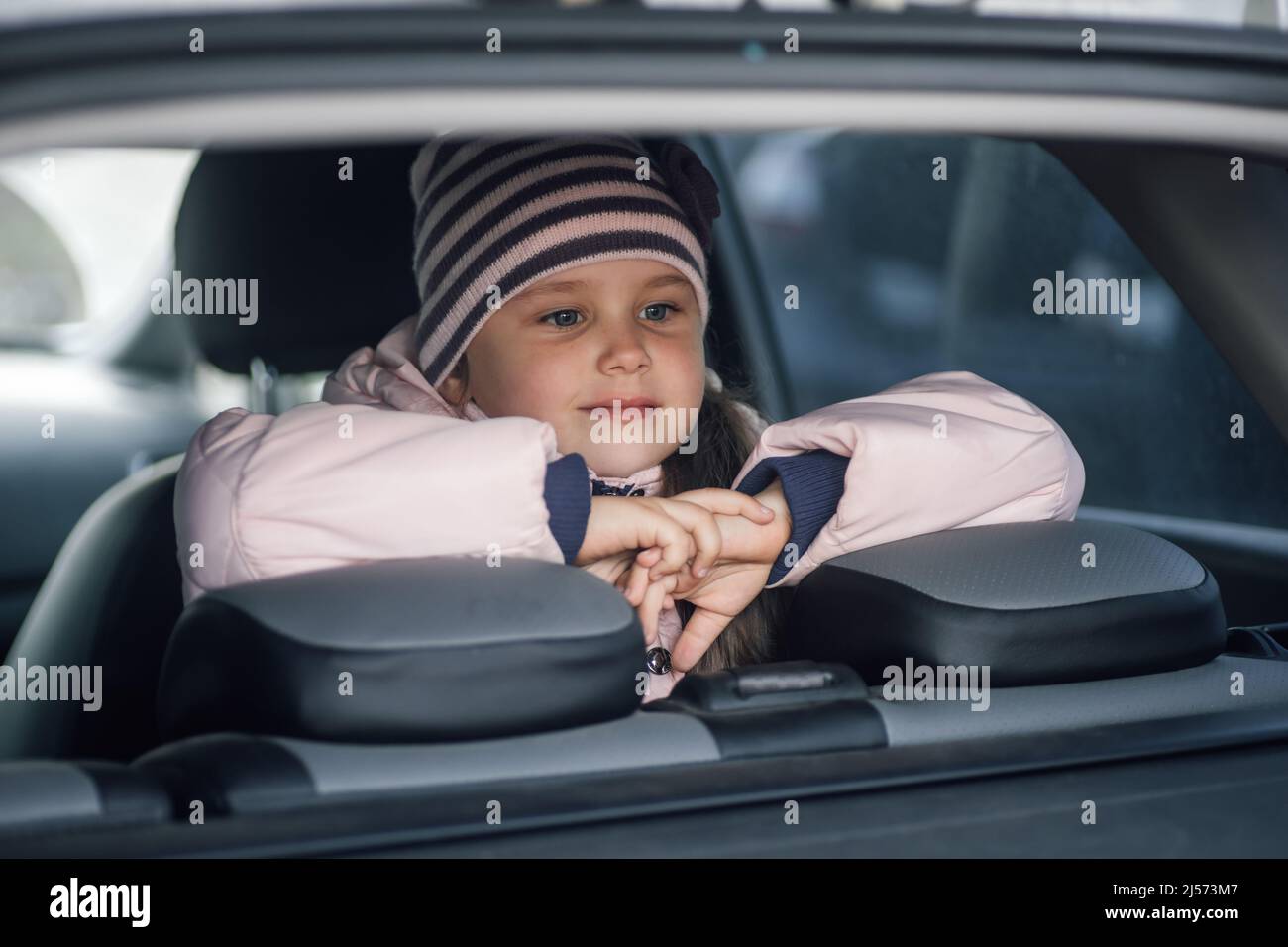 Das junge weibliche Kind sitzt hinten im Auto und schaut durch das Kofferraumfenster mit den Händen auf den Kopfteilen der Autositze, um irgendwo in Pink zu fahren Stockfoto