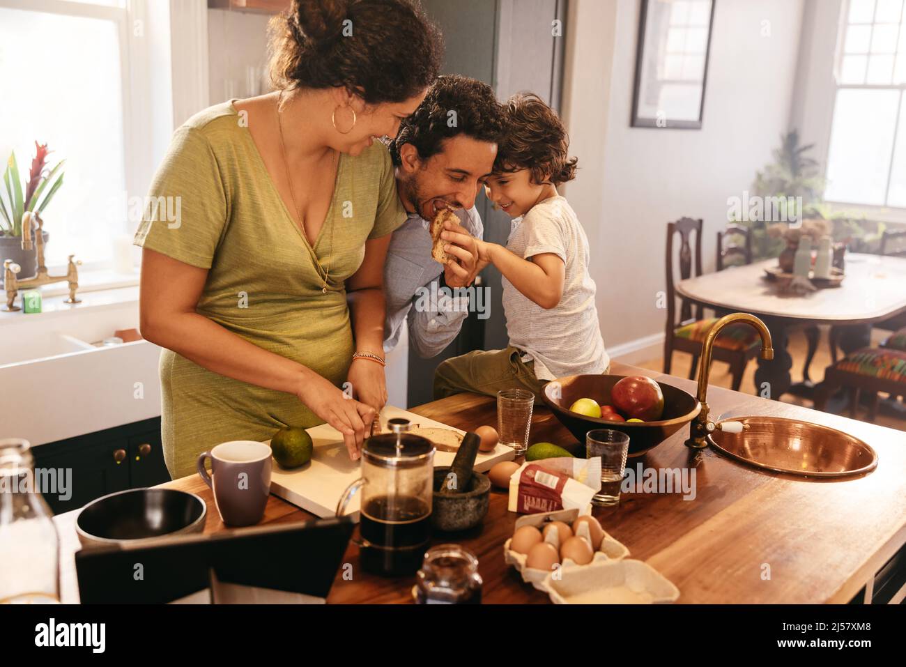 Dreiköpfige Familie, die eine gute Zeit in der Küche verbringt. Verspielter Vater, der ein Stück Brot isst, während seine Frau Frühstück macht. Junge Familie, die Spaß hat Stockfoto