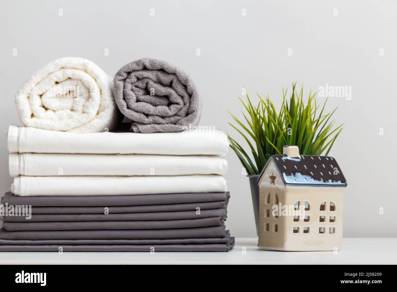 Ein Stapel grauer und weißer Bettwäsche, Bettwäsche und Frottee-Handtücher auf dem Tisch. Eine Topfpflanze und ein Spielzeug-Häuschen Stockfoto