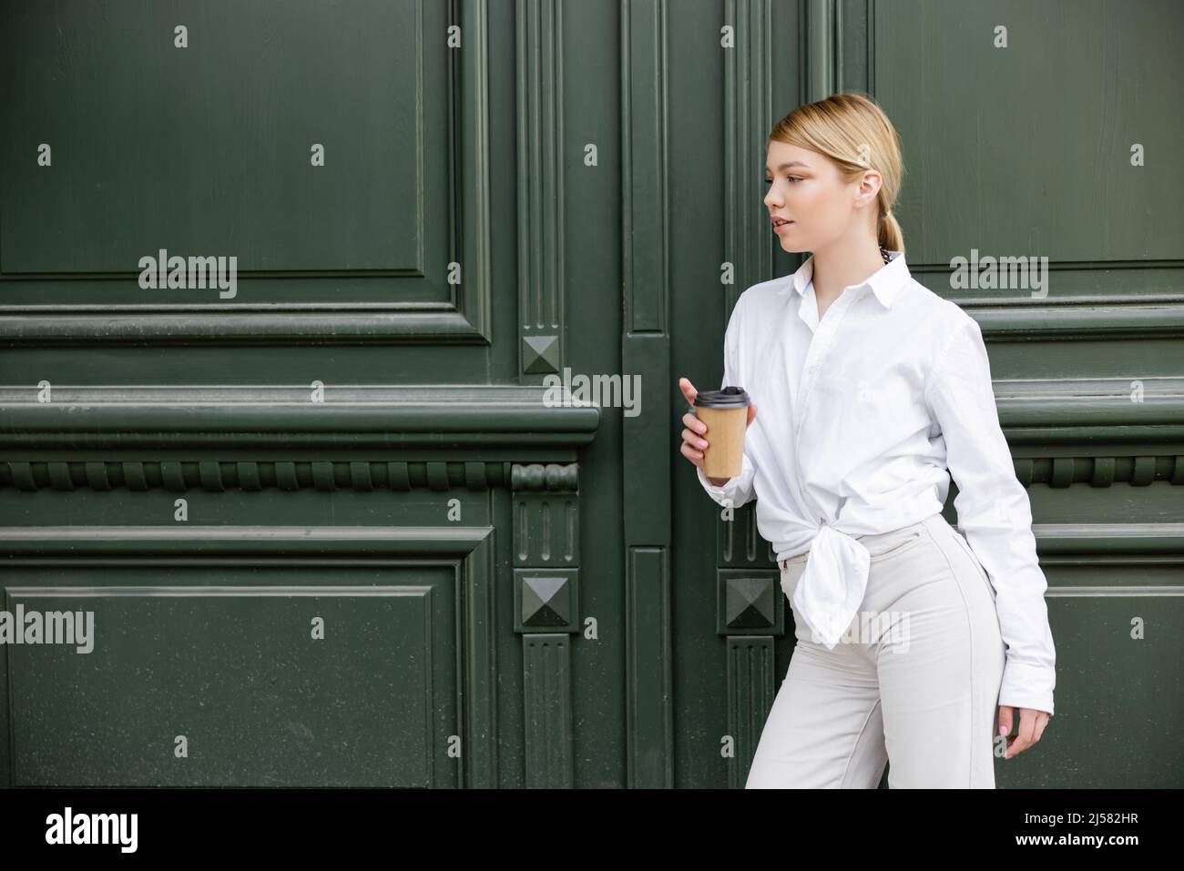 Hübsche Frau in weißem Hemd und Hose mit Pappbecher in der Nähe der grauen Wand Stockfoto