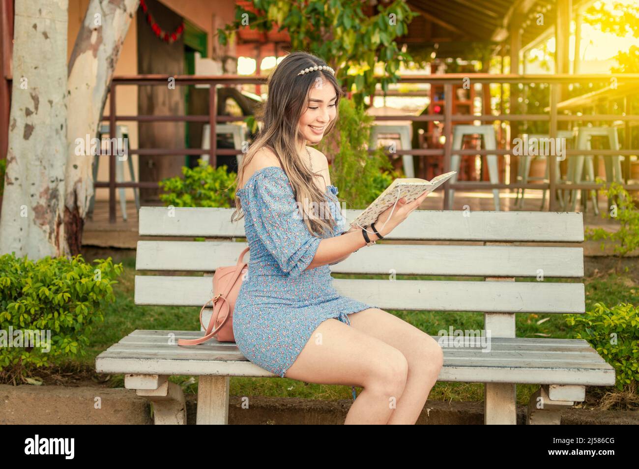 Ein süßes Mädchen, das auf einer Bank sitzt und ein Buch liest, ein hübsches junges lateinisches Mädchen, das ein Buch auf einer Bank liest Stockfoto