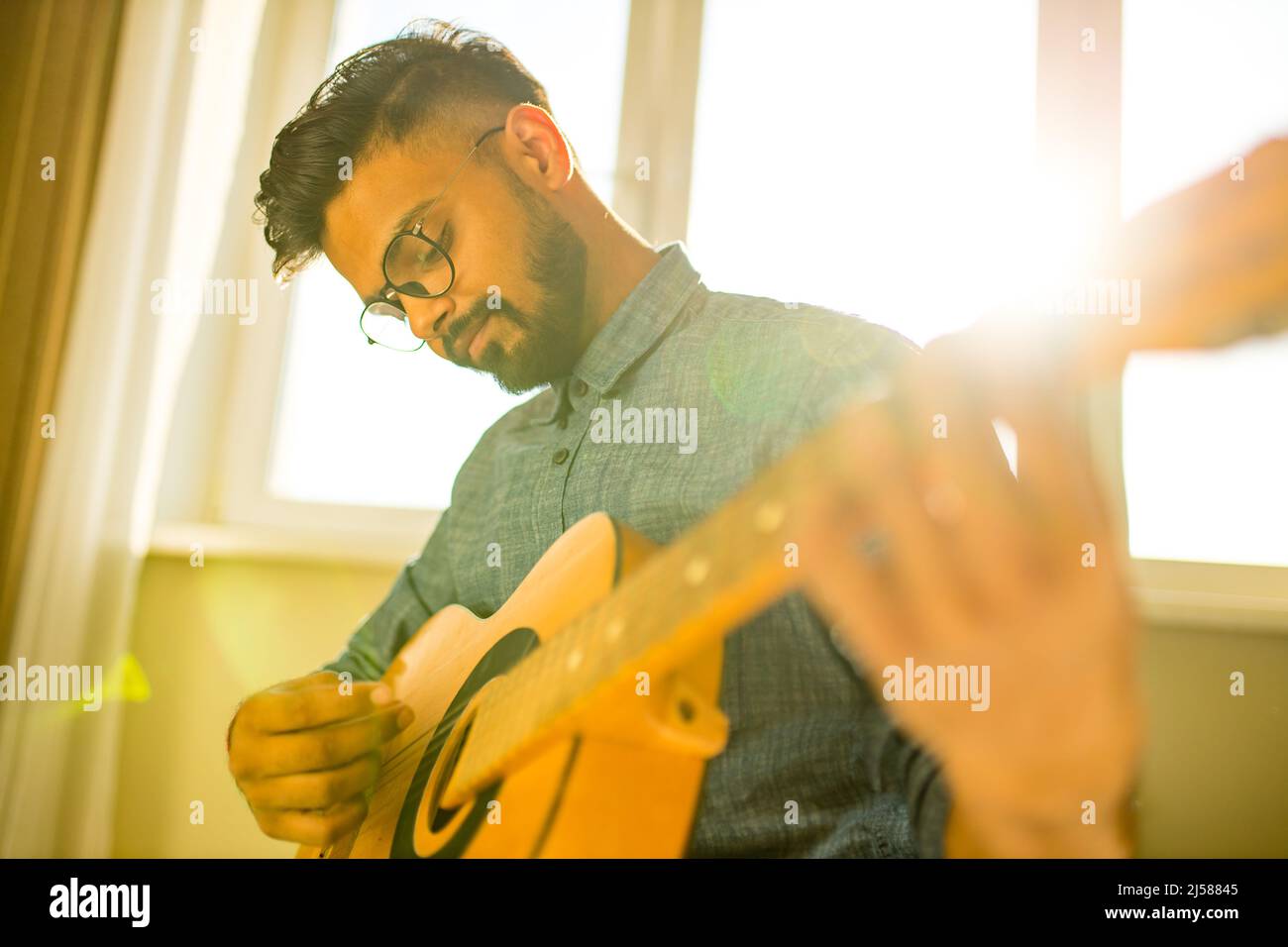 Fröhlicher, hübscher junger Mann, der Gitarre spielt und lächelt in einer neuen, gemütlichen Wohnung Stockfoto