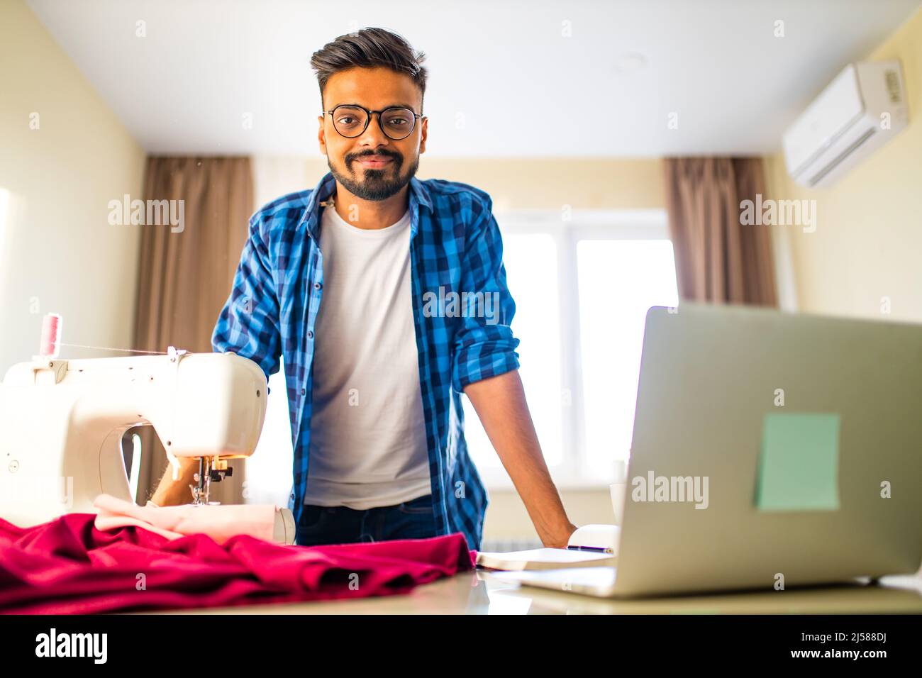 indische Schneider asiatische Mann Designer nähen Kleidung auf Nähmaschine neben Laptop Wohnung Arbeitsplatz Stockfoto