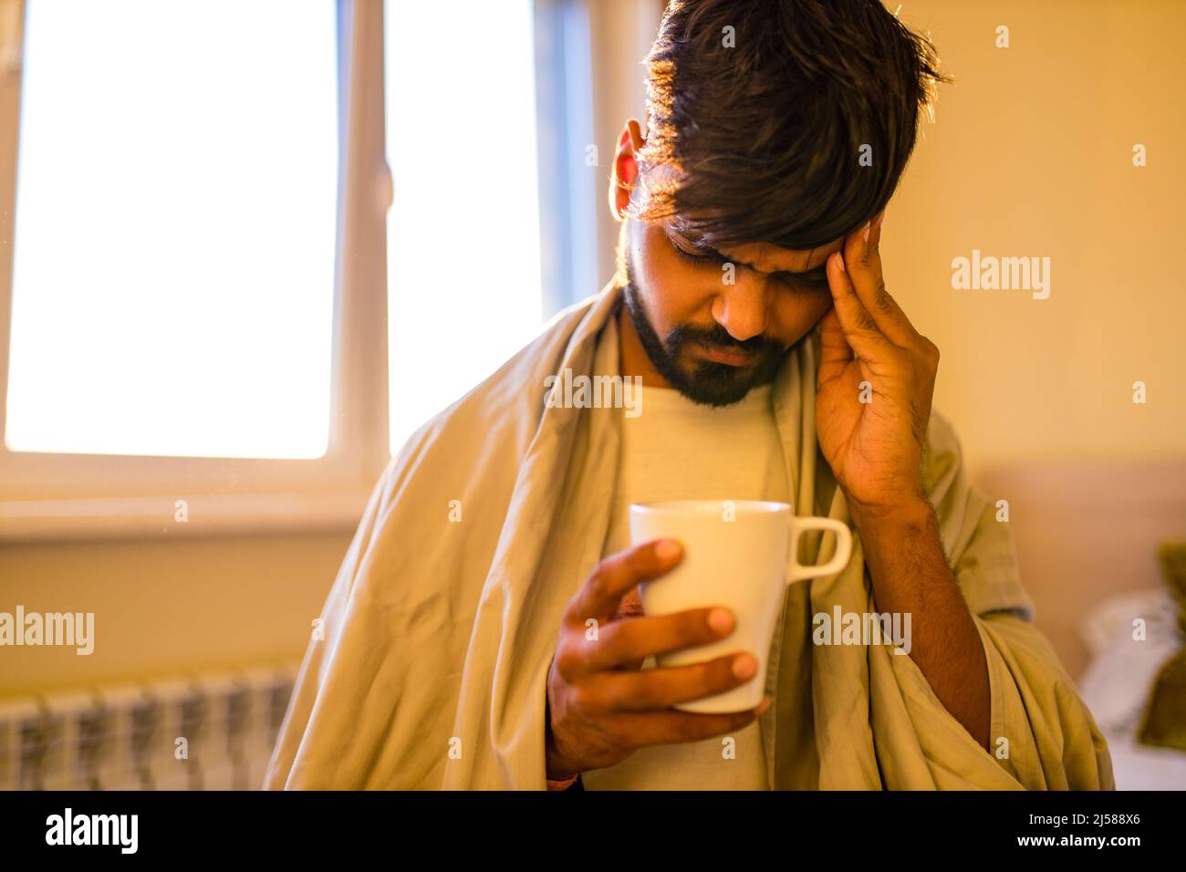 arabischer indischer Mann trinkt heißen Zitrone Dinder Masala Tee im Schlafzimmer am Morgen, während das Gefühl kalt mit einer Decke bedeckt Stockfoto