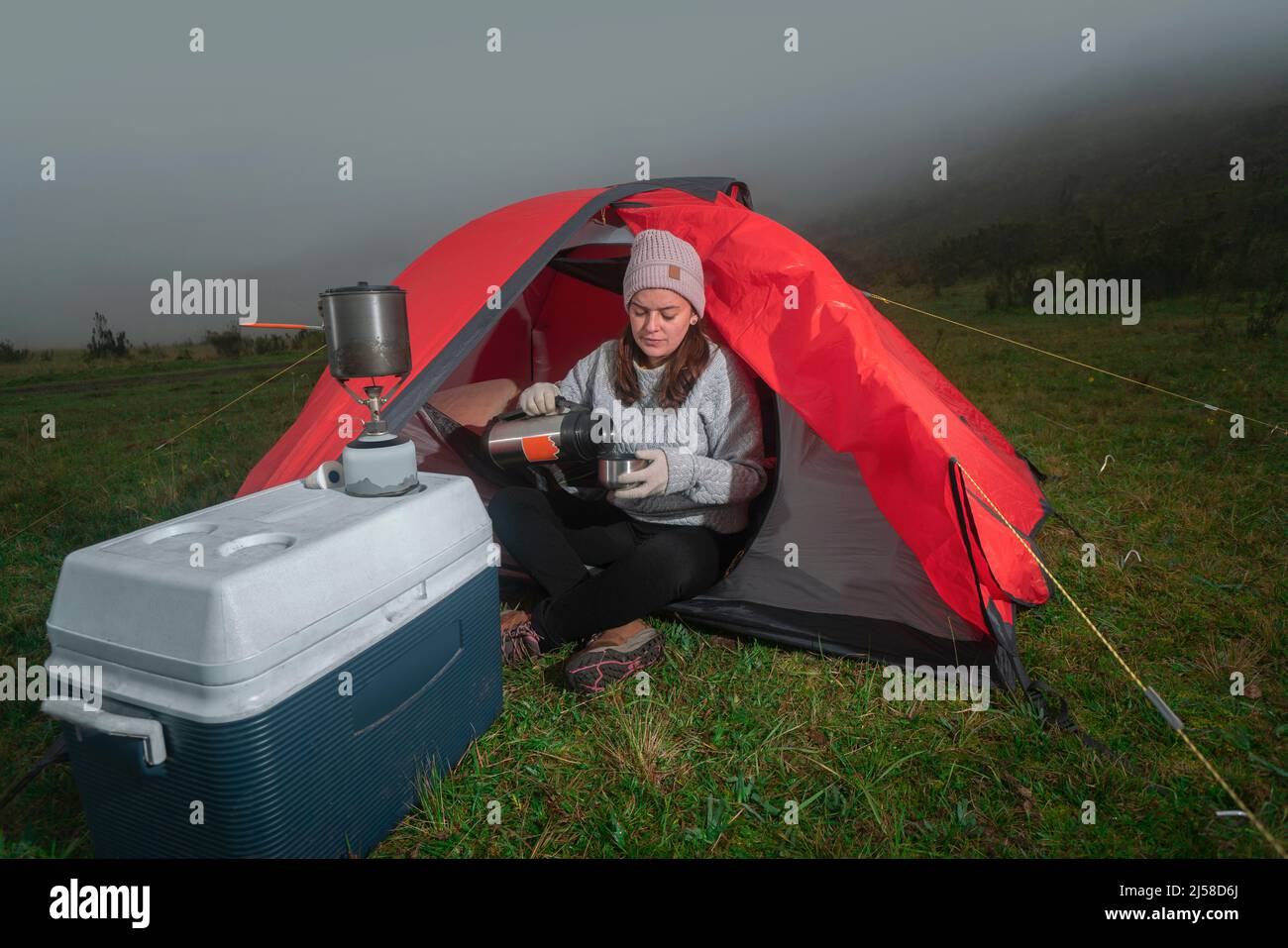 Schöne Lateinerin, die allein vor ihrem roten Zelt inmitten eines grünen Feldes sitzt und Tee aus ihrer Thermoskanne neben einem Kühler in einer bewölkten Sonne trinkt Stockfoto