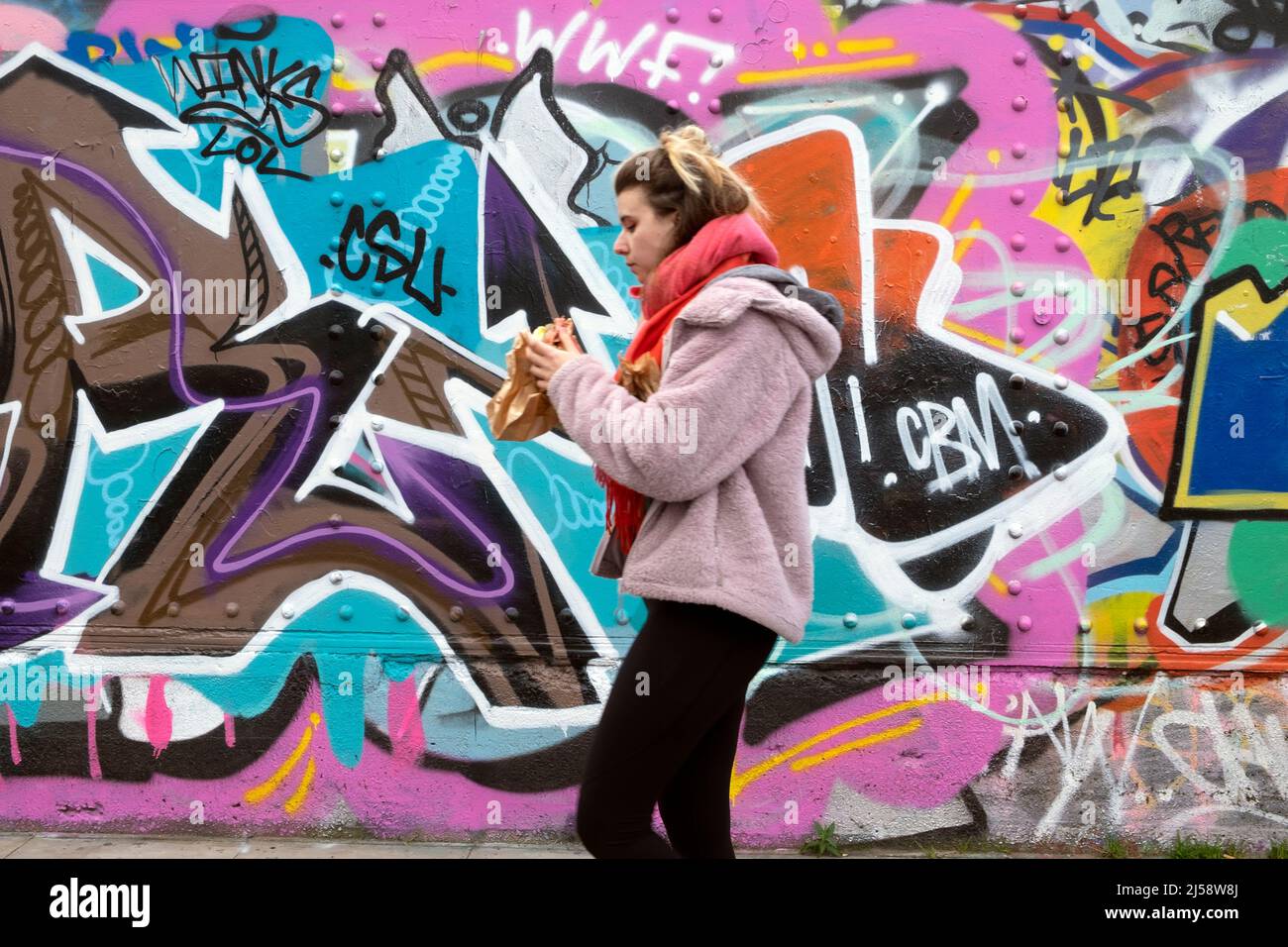 Person Frau, die an einem Wandgemälde auf dem Horten in der Brick Lane vorbei geht Shoreditch, der Bagel isst April 2022 London England Großbritannien KATHY DEWITT Stockfoto