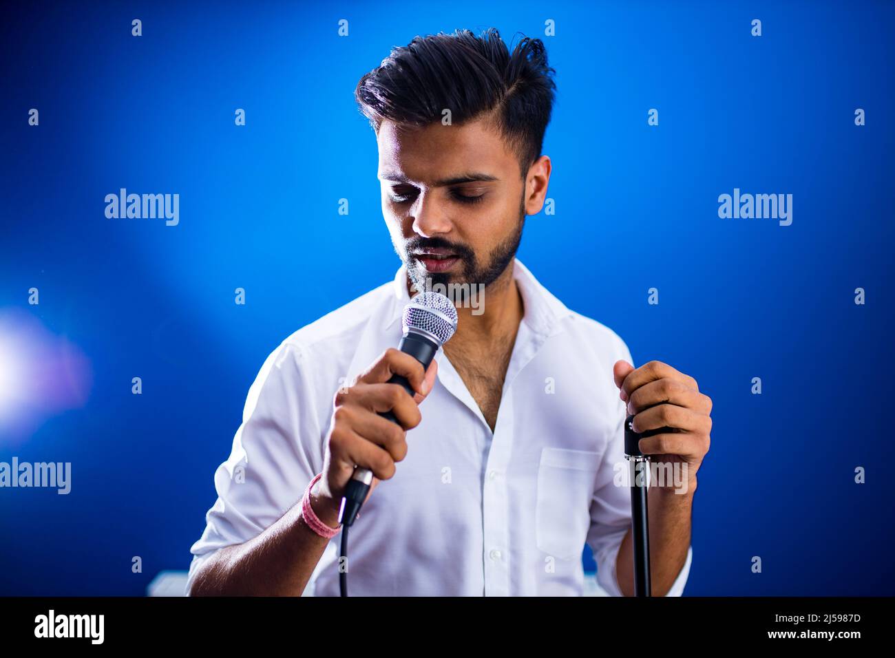 indischer Mann im weißen Hemd mit Bart, der im Aufnahmestudio auf blauem Hintergrund singt Stockfoto