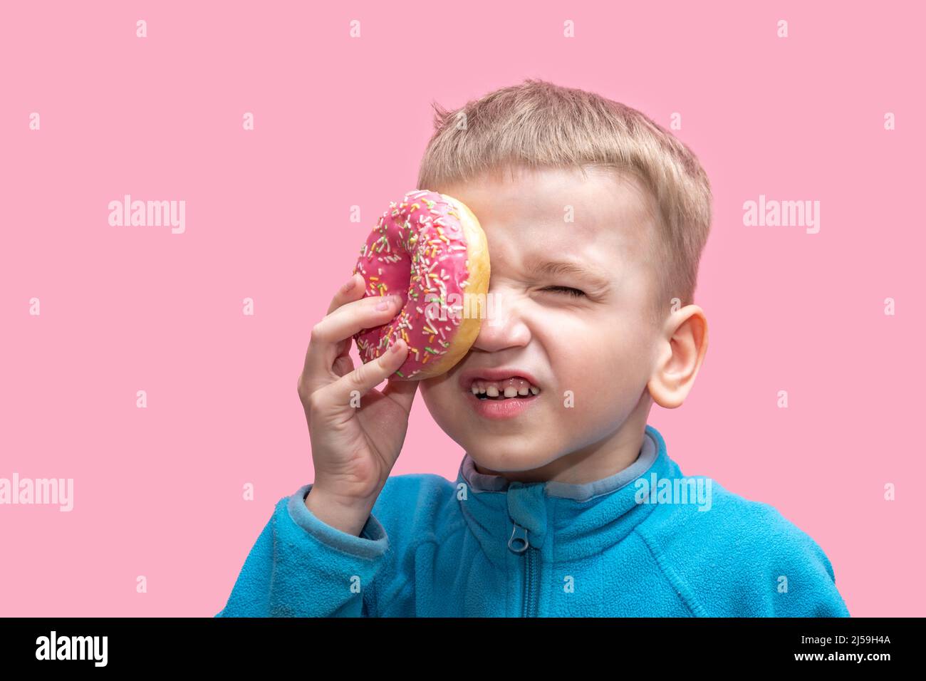 Ein süßer, lustiger Junge in einem blauen Pullover hält einen leuchtend rosa Donut in der Nähe seines Auges und schielt auf einem rosa Hintergrund. Fröhliches Kindheitskonzept. Süßes Essen für Stockfoto